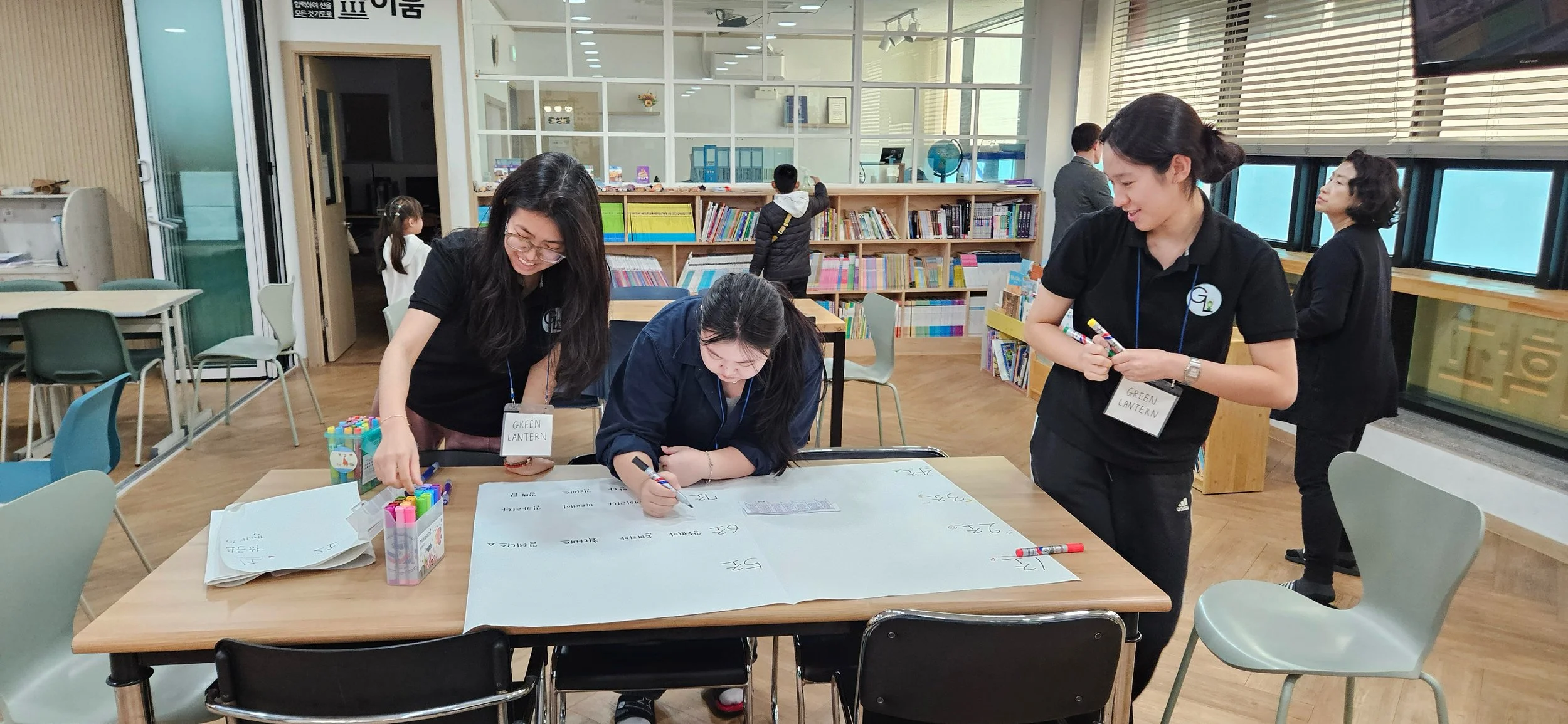 Group of women working together at a table in a library, writing on a large sheet of paper with markers, with bookshelves and other children in the background.