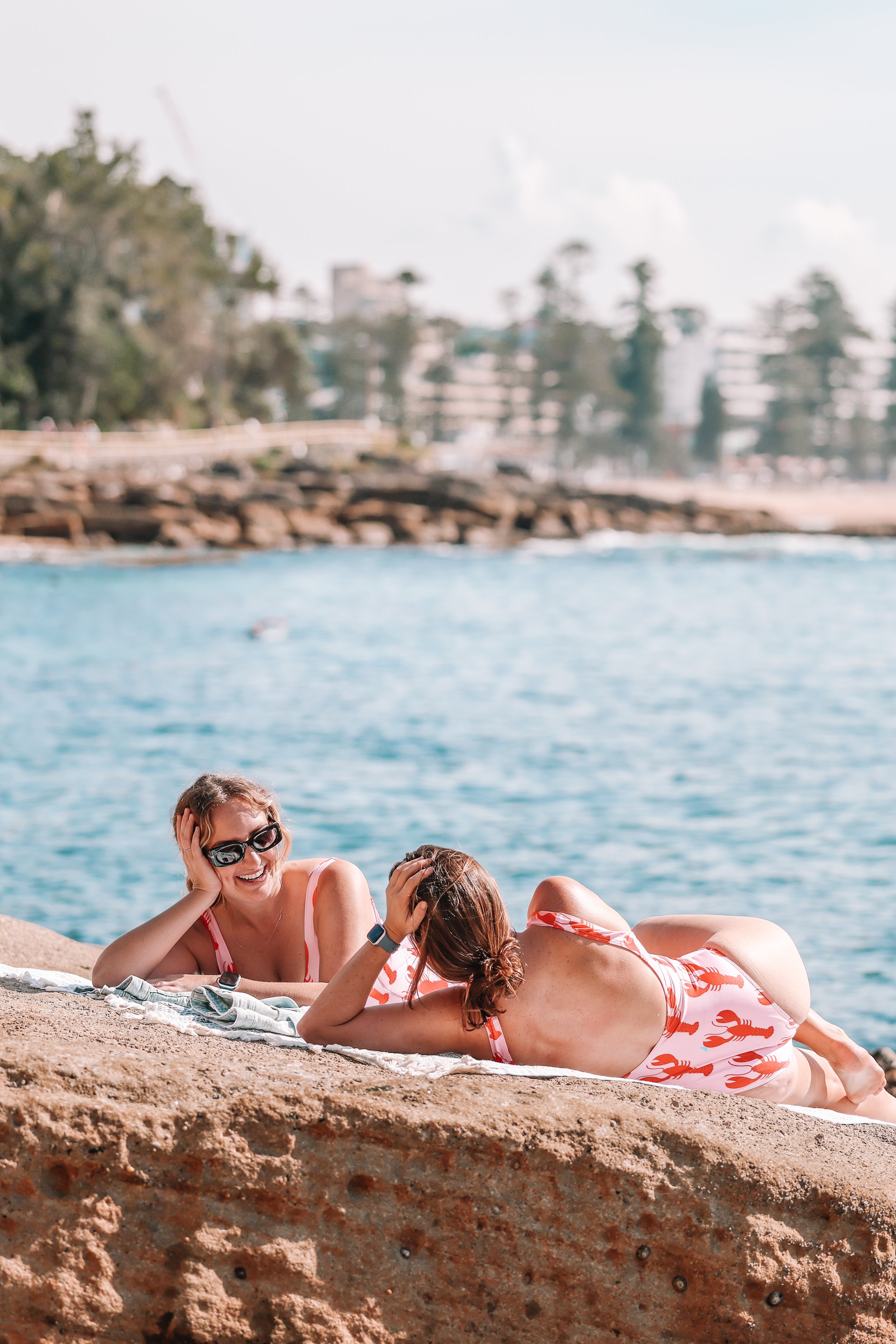 Two women are lying on a towel on the beach, relaxing and laughing together. They are wearing swimsuits and sunglasses, with trees and buildings in the background near the water.