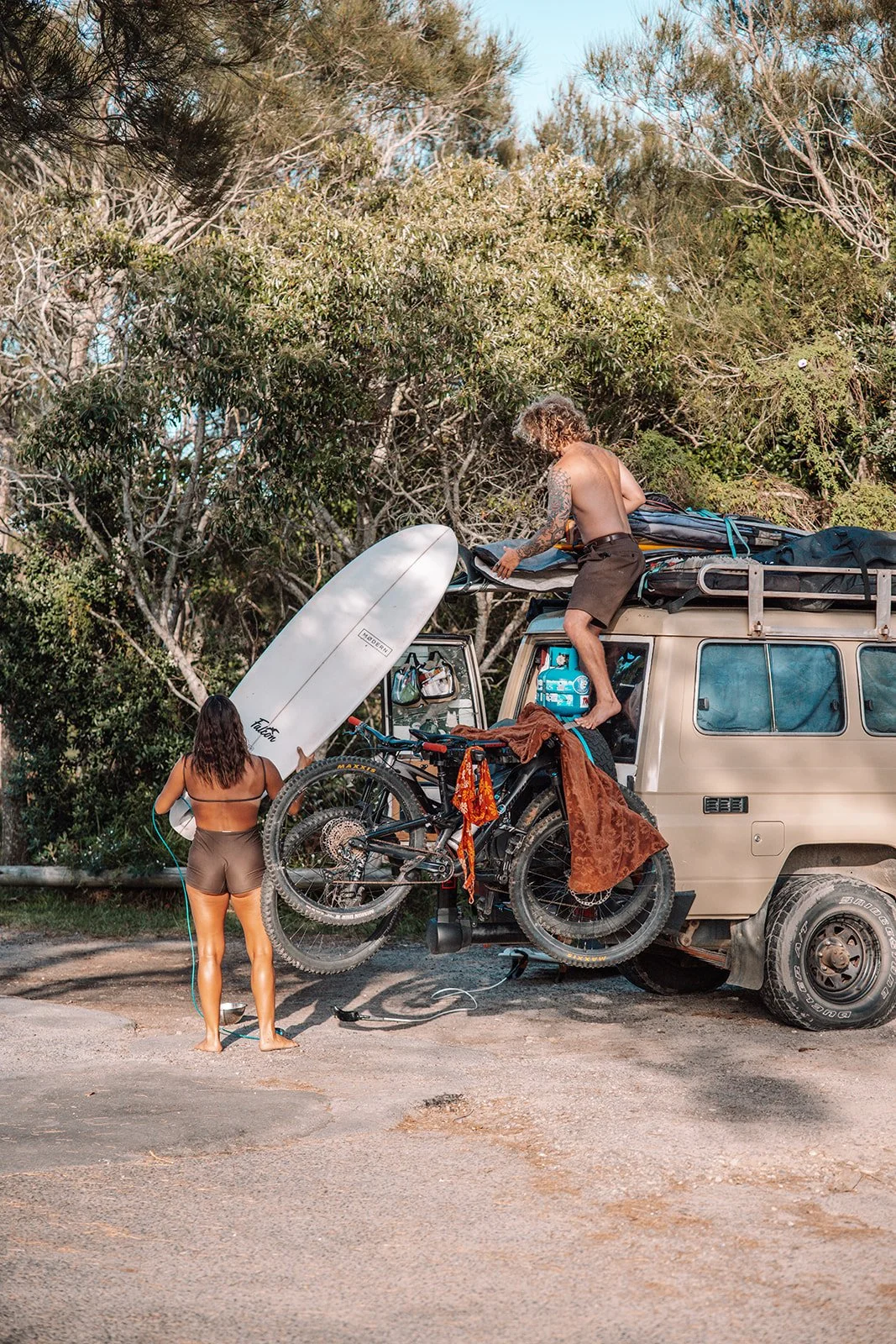 Two people preparing surfboards, a bicycle, and gear on top of a beige van in a wooded outdoor area.