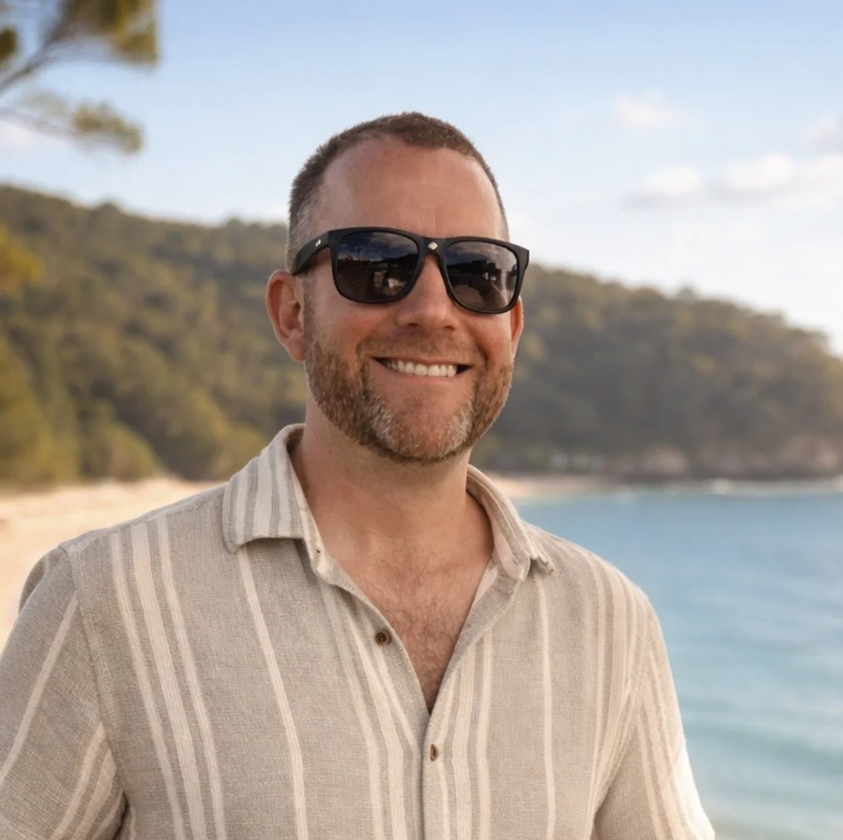 Smiling man wearing sunglasses and a striped beige shirt outdoors on a beach with trees and water in the background.