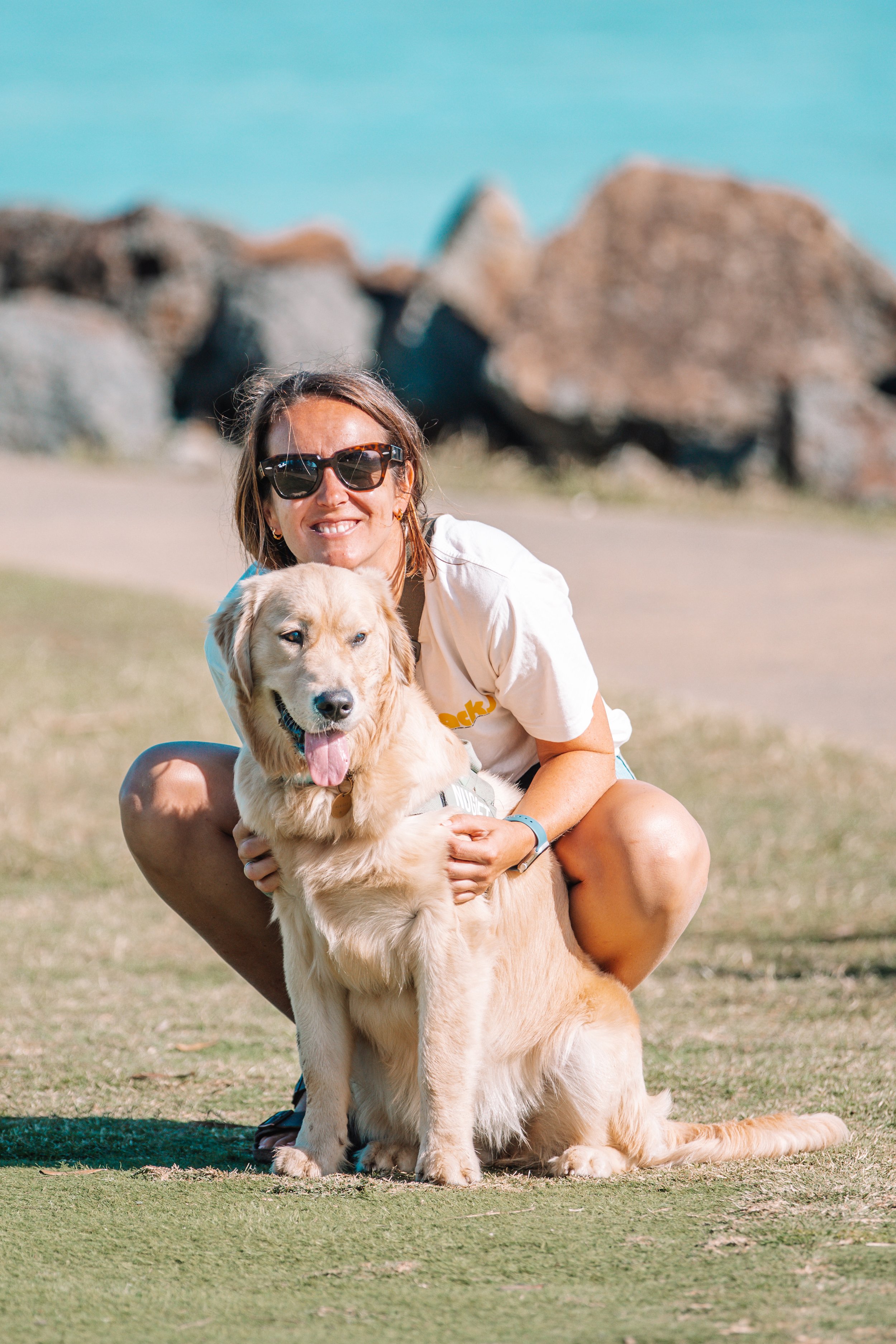 A woman with sunglasses smiling while hugging a golden retriever dog on a grassy field with rocks and clear blue sky in the background.