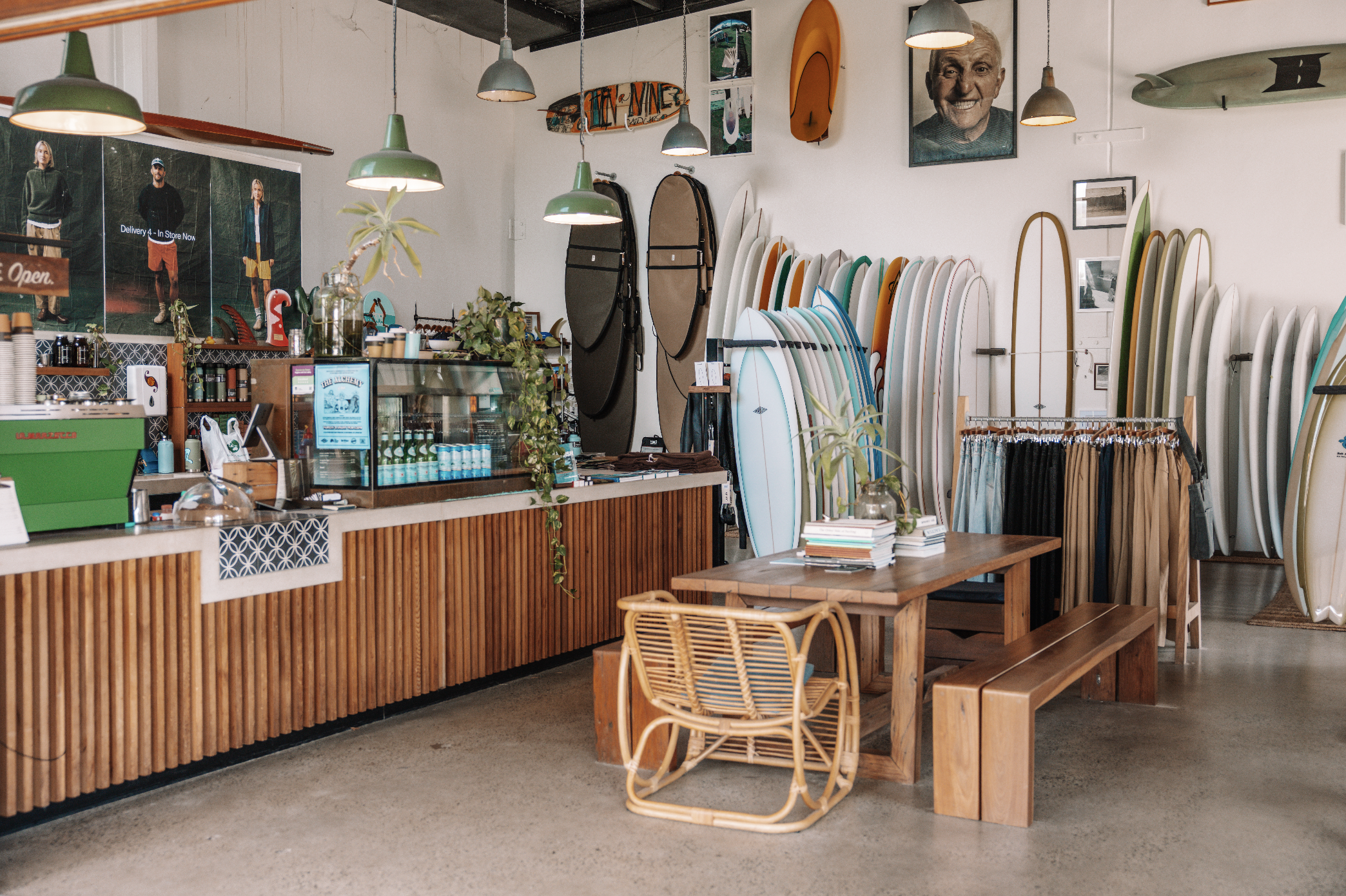 Interior of a surf shop with surfboards lined up on the wall, a wooden counter with plants and merchandise, and a seating area with wooden tables and chairs. Various colorful surfboards are displayed, and framed photos and artwork adorn the walls.