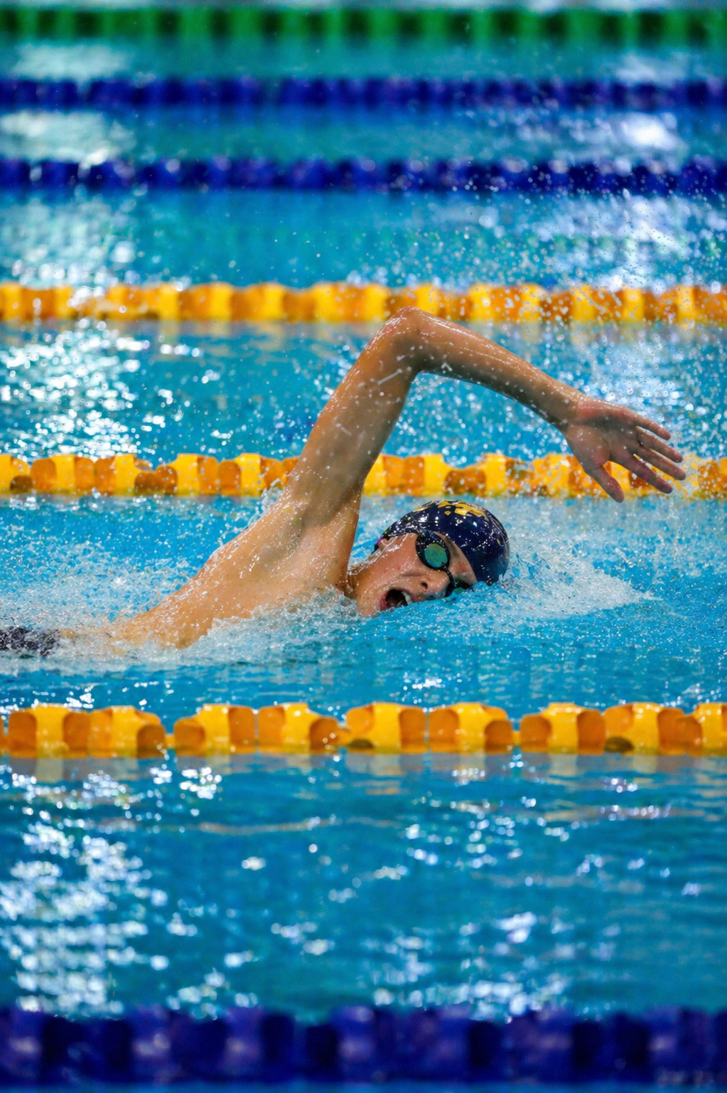 A swimmer wearing goggles is seen mid-stroke in a swimming pool during a race, with yellow lane dividers visible.
