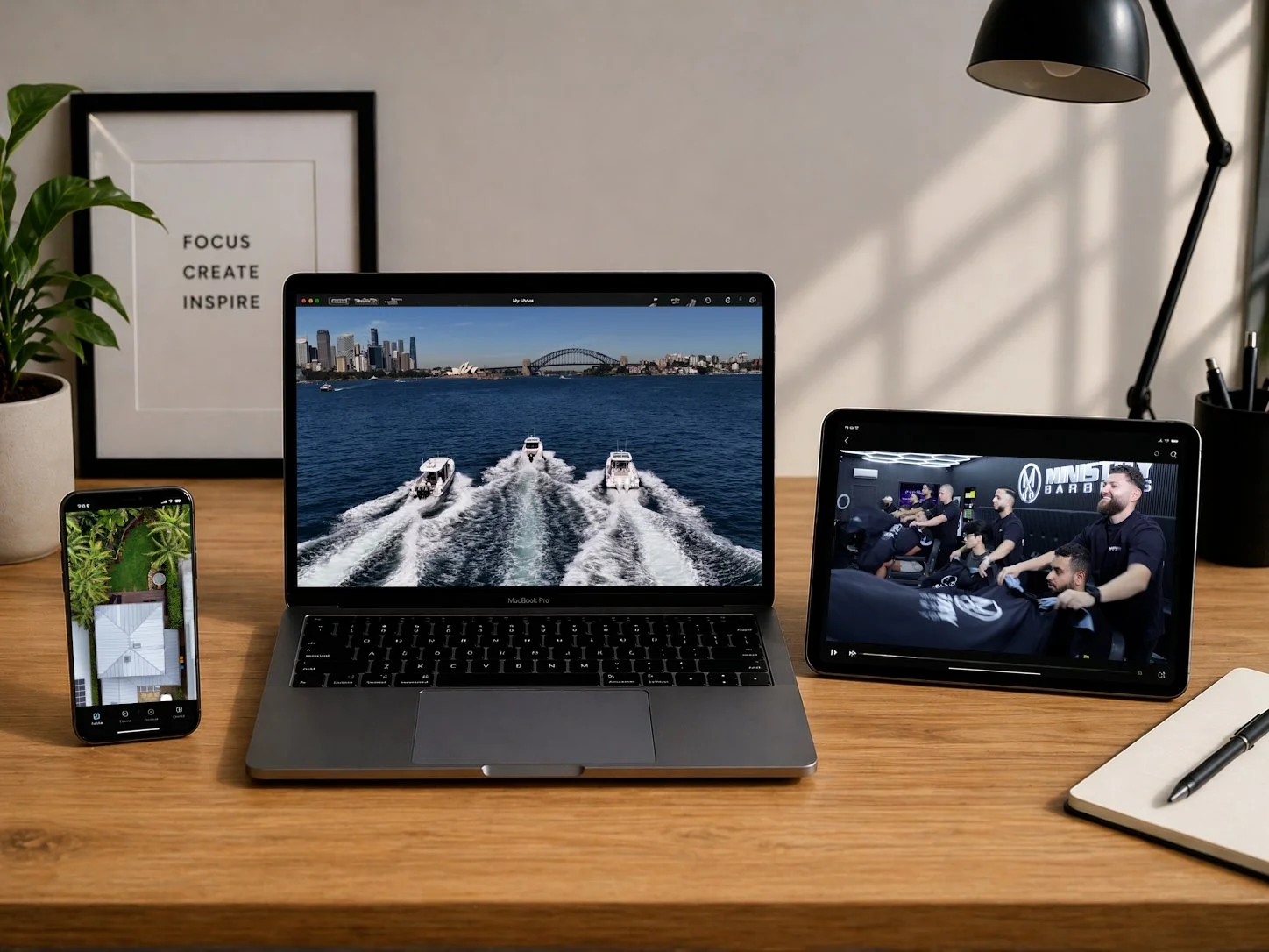 A workspace with a MacBook Pro displaying a photo of boats on a waterway, an iPhone with an aerial view of a house surrounded by trees, and a tablet showing people at a gym, on a wooden desk with a potted plant, a framed motivational quote, a black desk lamp, a pen, and a notebook.
