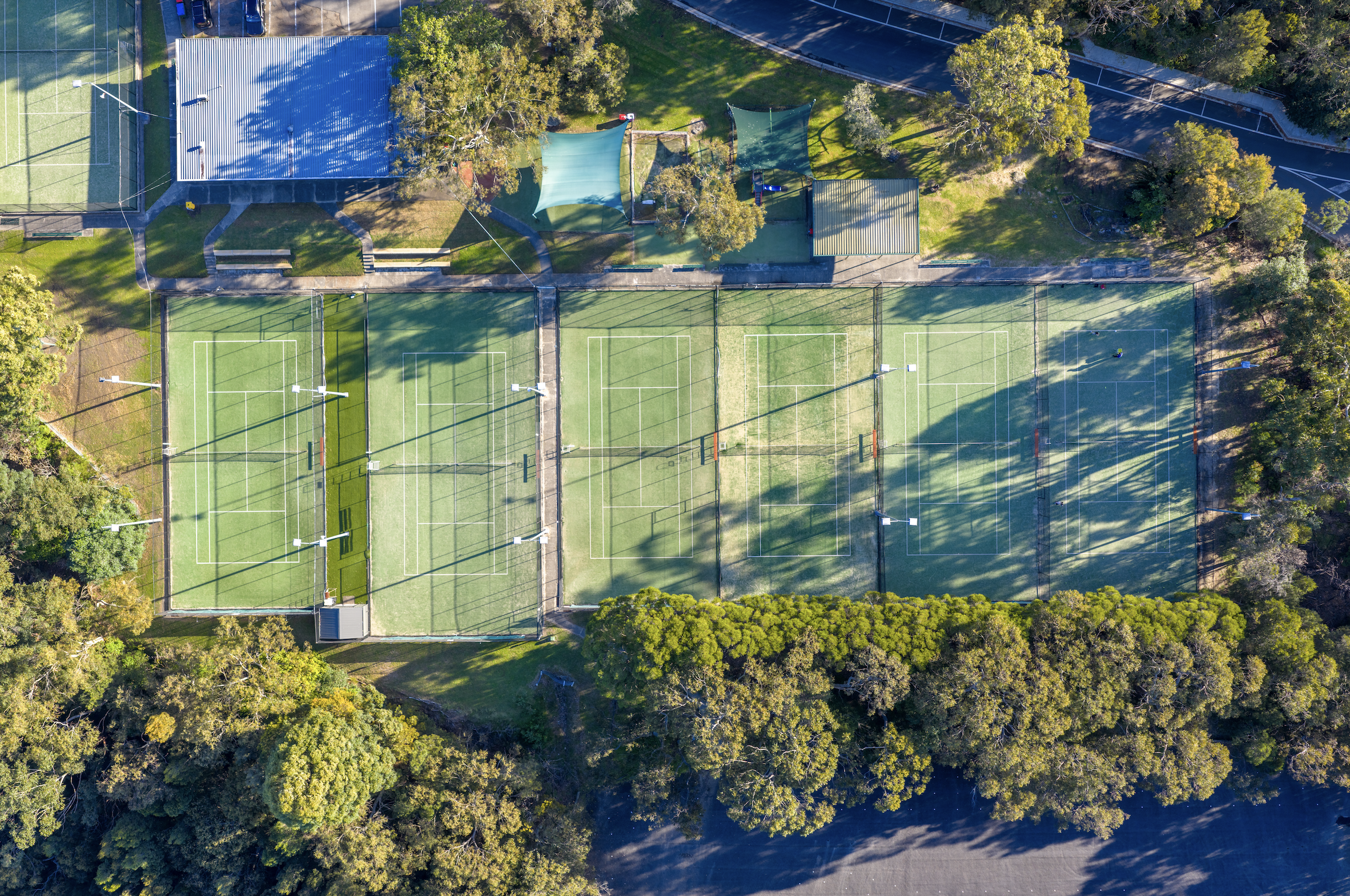 An aerial view of a sports facility surrounded by trees, showing two tennis courts, a playground, and a building with a blue roof visible in the upper left corner.