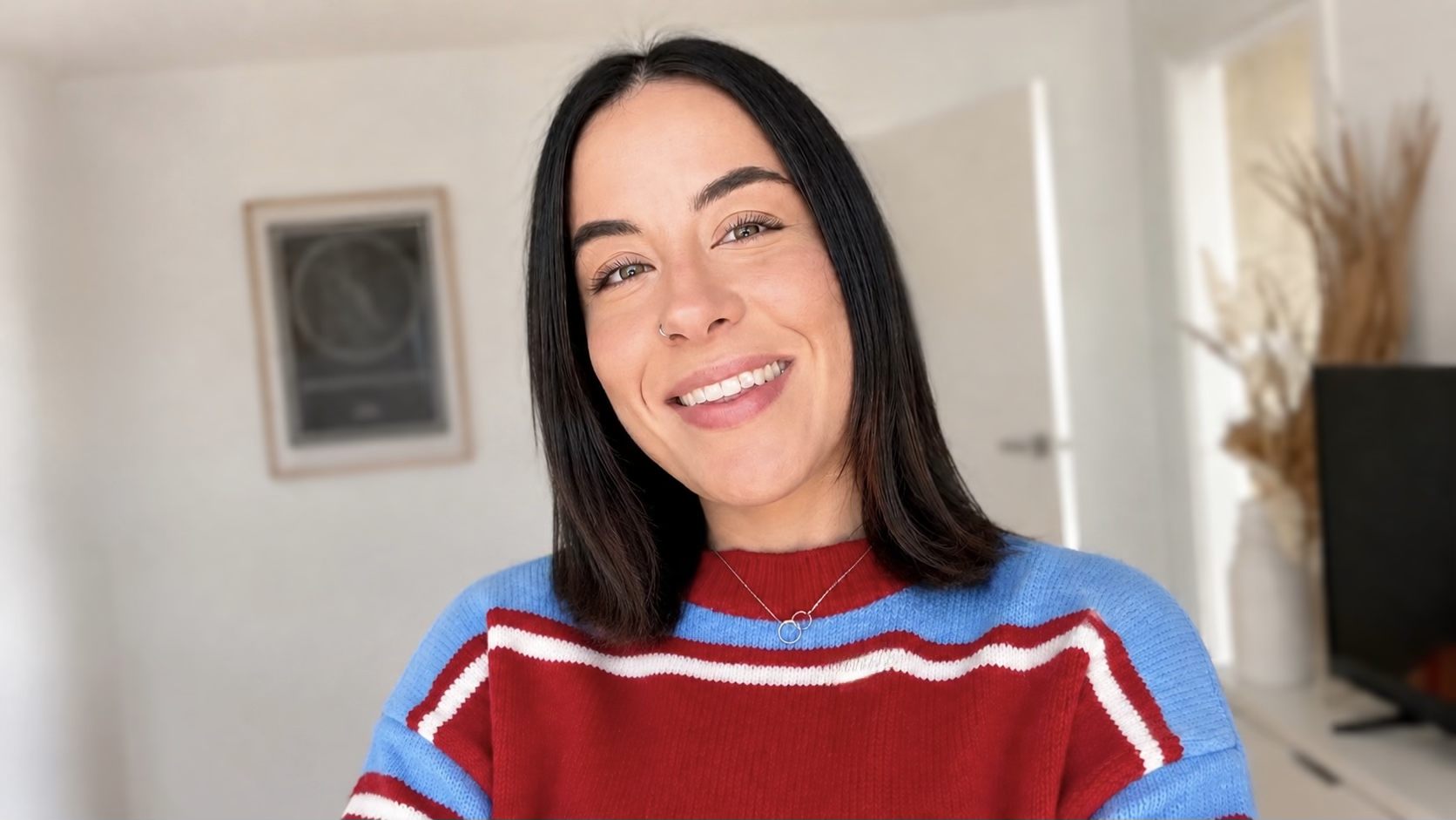A smiling woman with shoulder-length dark hair, wearing a red and blue striped sweater, stands in a well-lit room with a white door, a TV, and a tall vase with dried decorative branches in the background.