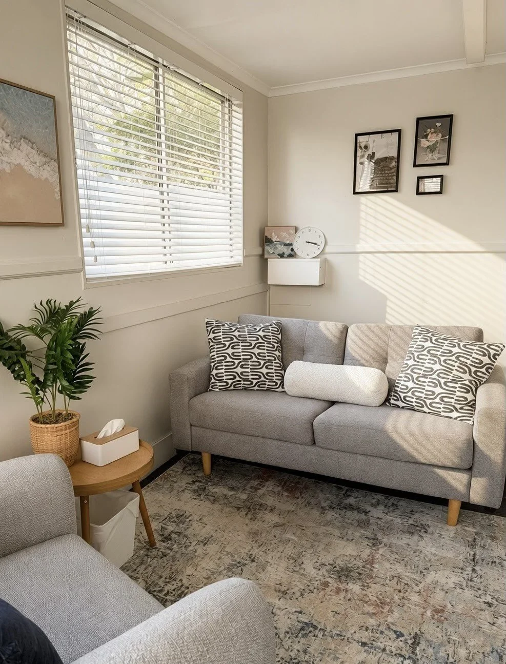 A cozy room with a beige sofa, decorative pillows with black and white patterns, a wooden side table with a potted plant and tissue box, a large window with white blinds casting light, framed wall art, a white wall clock, and a textured rug.