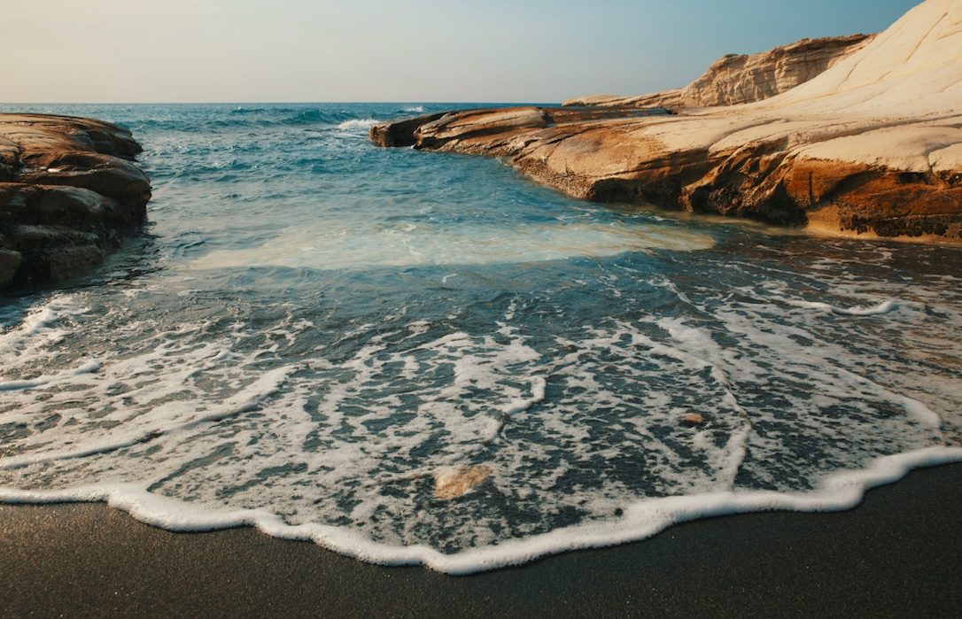 A beach scene with waves washing over dark sand, rocky formations on the right side, and the ocean extending to the horizon under a clear sky.