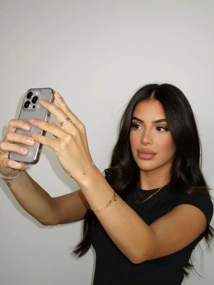 Une femme prenant un selfie avec un smartphone, portant des bijoux, fond uni.