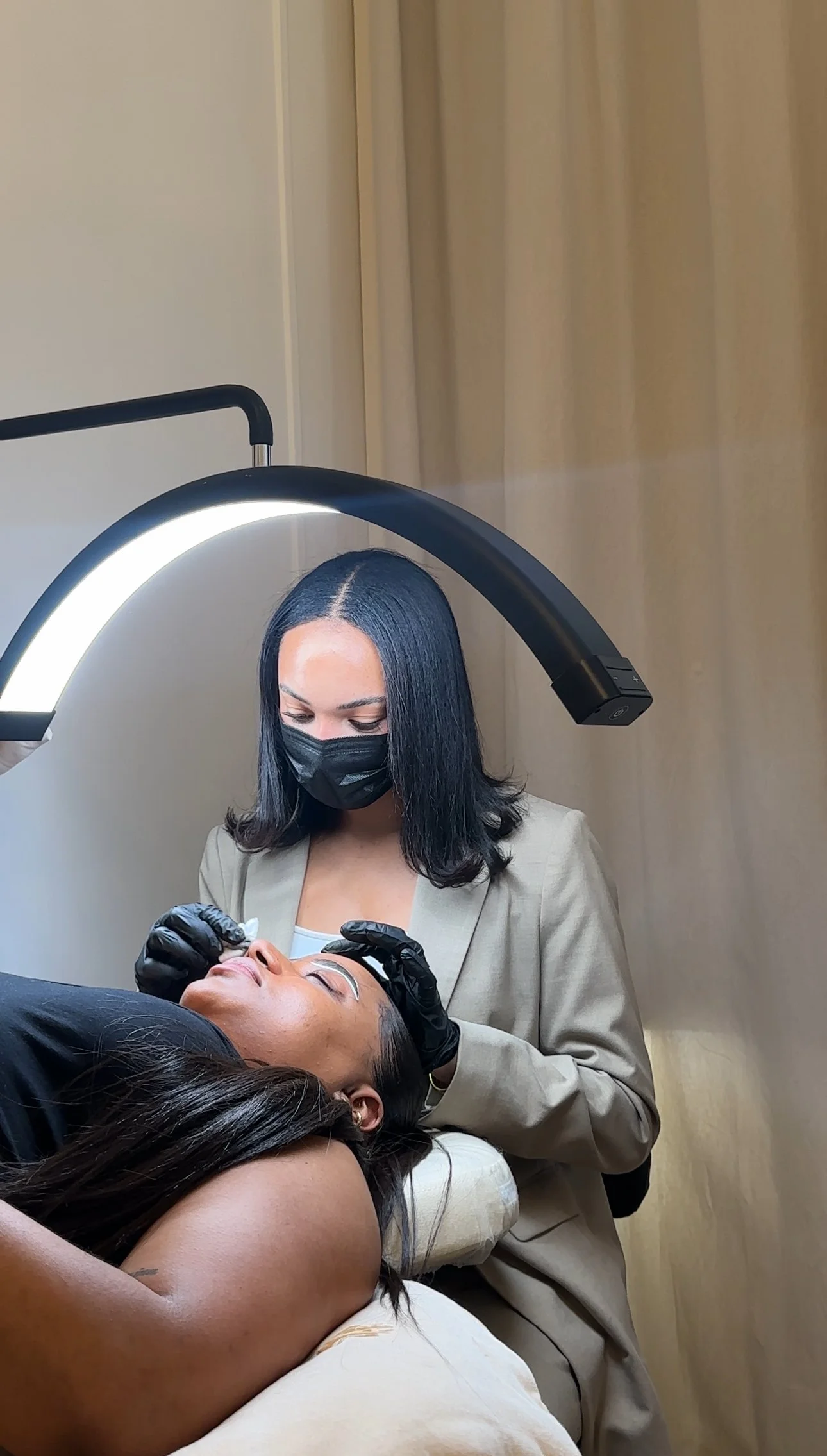 Une esthéticienne portant un masque applique un soin sur le visage d'une cliente allongée, éclairée par une lampe circulaire, dans un salon de beauté.