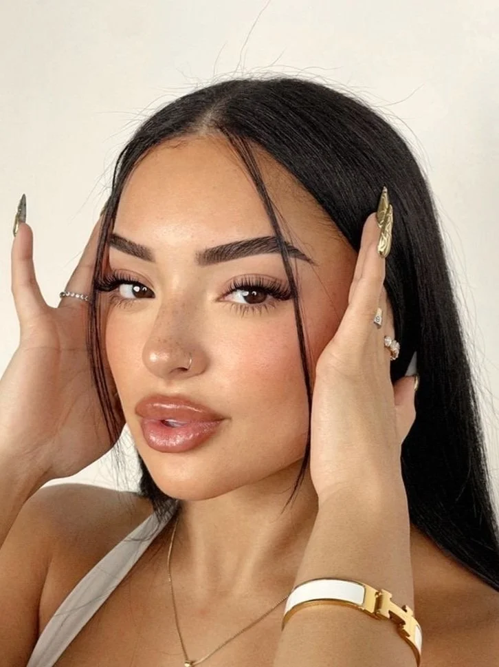 Une femme avec des cheveux foncés et des ongles longs, portant des bijoux, pose avec ses mains sur son visage.