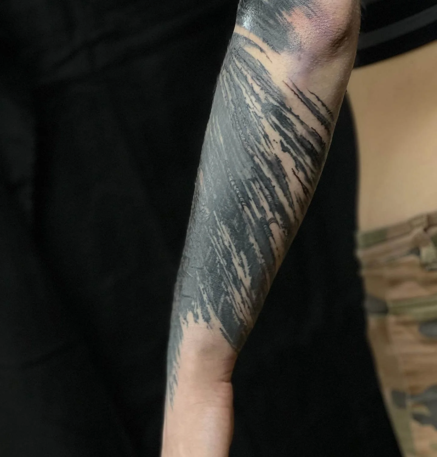 Black and grey abstract landscape tattoo featuring mountains, trees and layered sky elements on the arm.