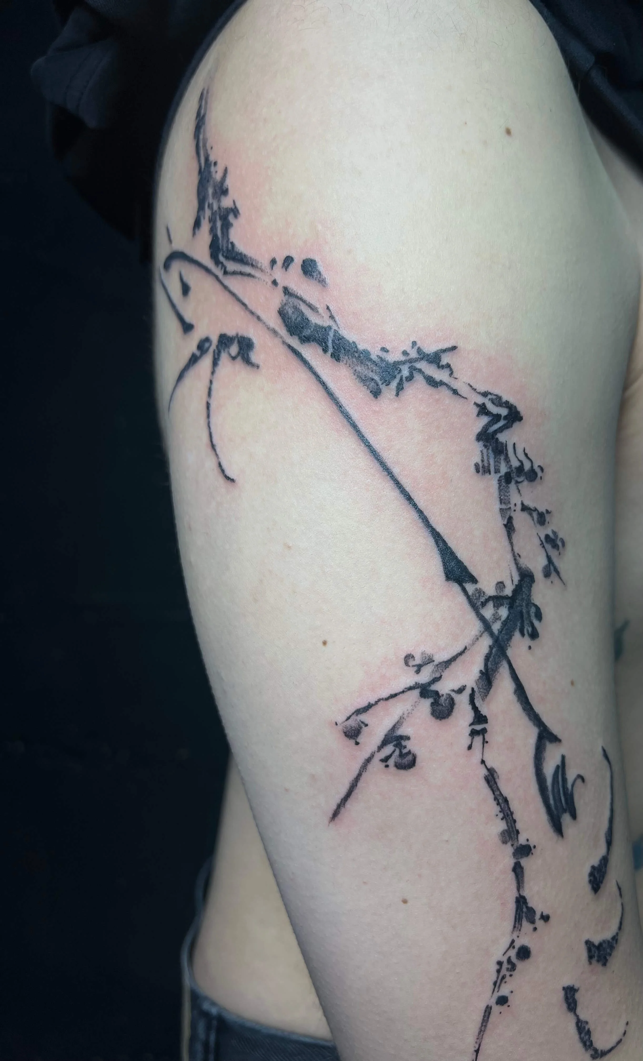 A black and gray outline tattoo of a delicate, abstract branch with small leaves and berries on a person's upper arm.