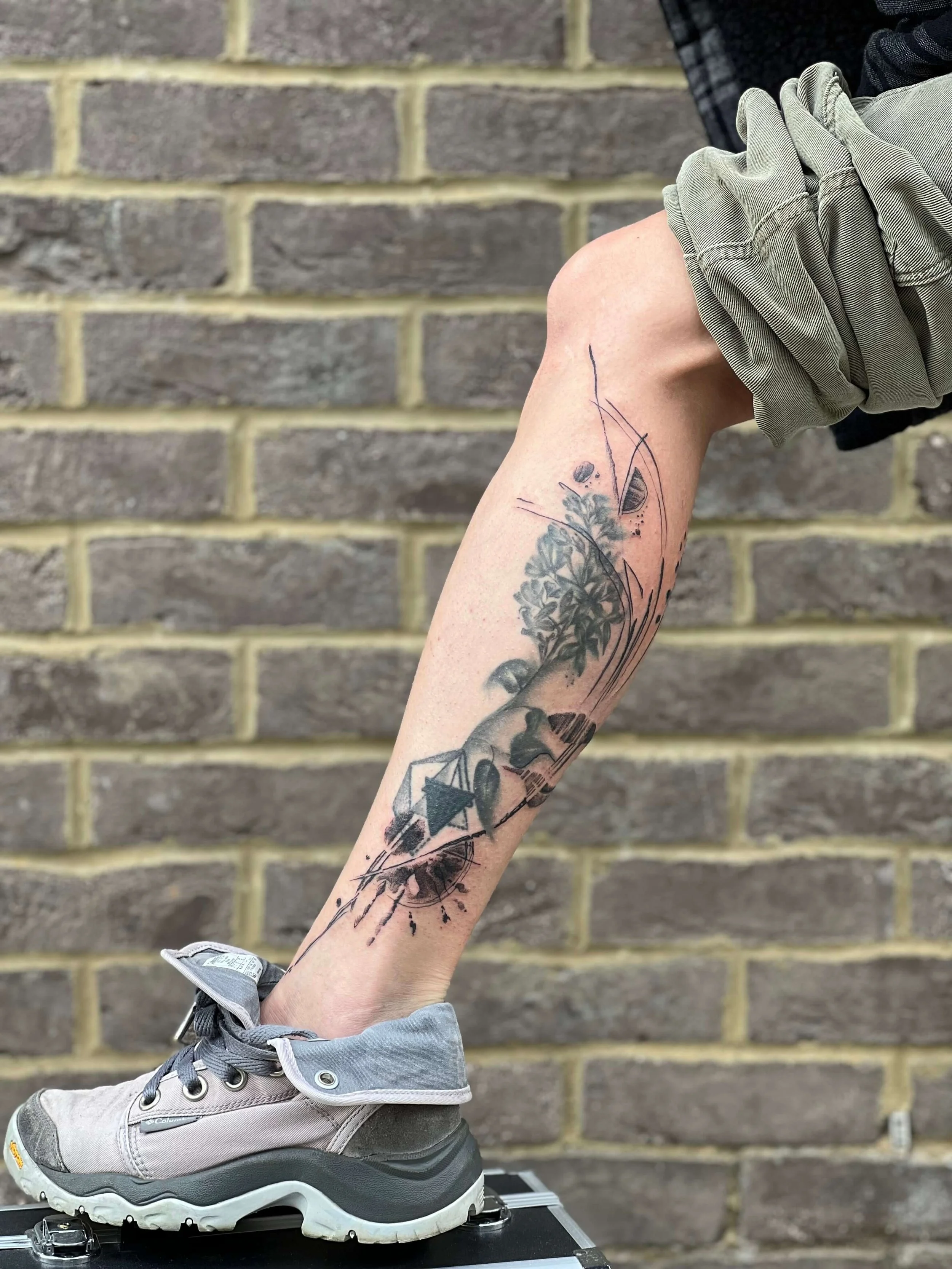 Abstract blackwork calf tattoo combining geometric structure, organic tree forms, and expressive ink splatter.