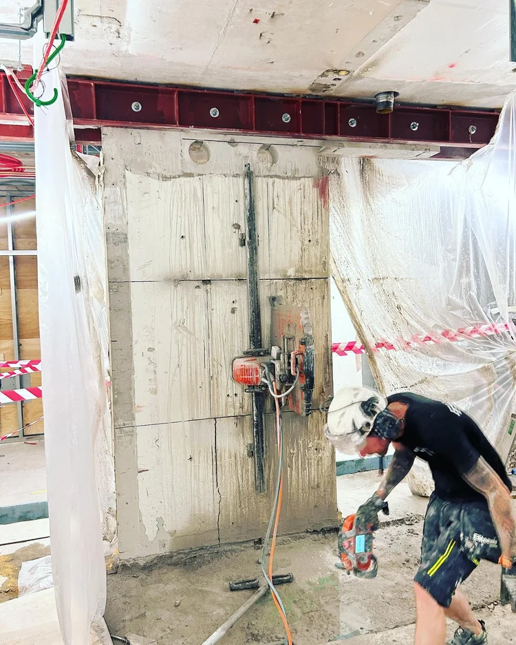 Byggarbete på en byggarbetsplats, där en person arbetar med borrutrustning mot en betongvägg. Skyddsplast hänger på sidorna och stålkonstruktioner syns ovanför. Personen bär skyddshjälm och byggkläder.