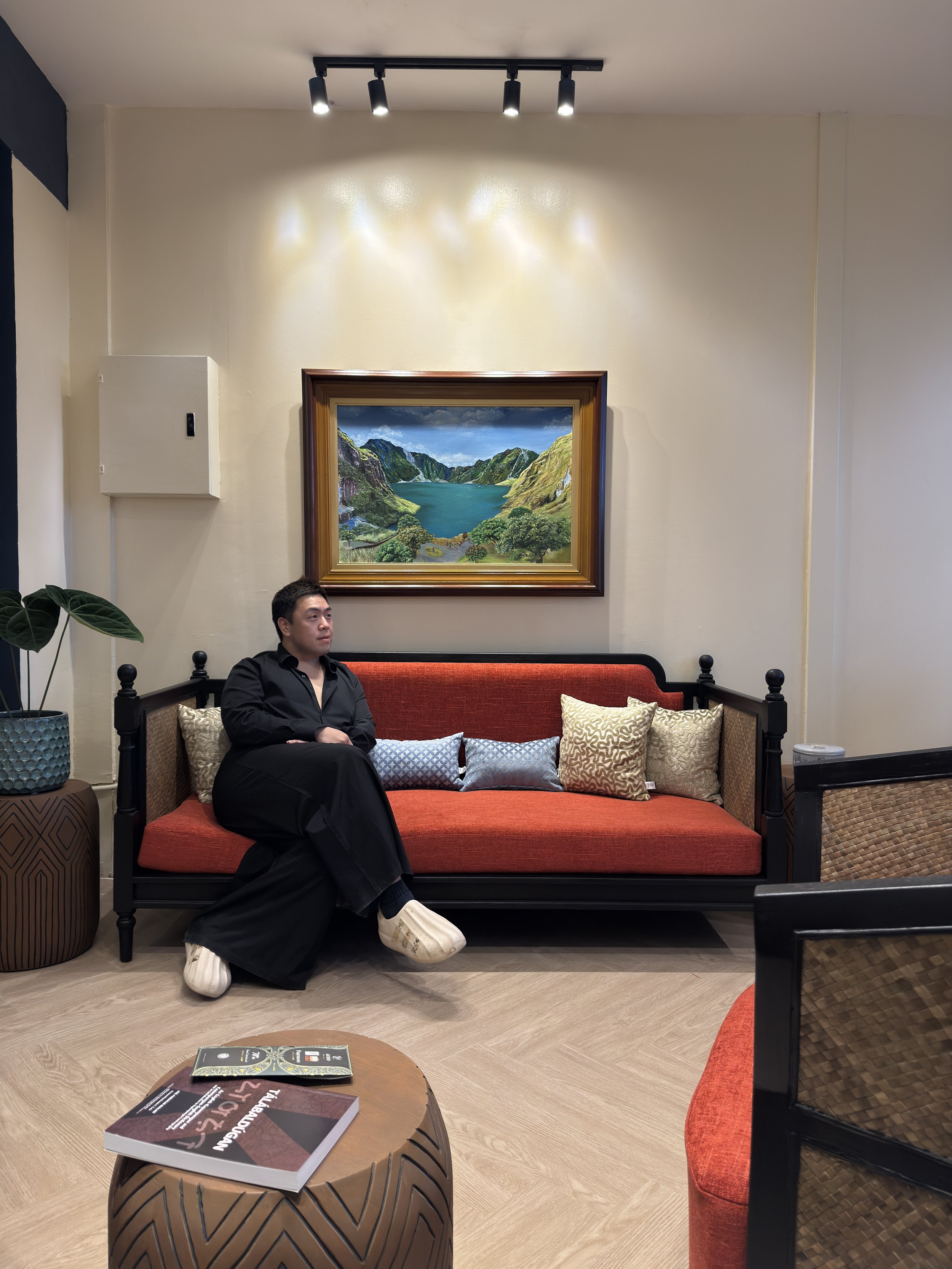 A man in black clothing sitting on a red sofa with seven decorative pillows, in a room with cream-colored walls. There is a framed landscape painting above him, a plant on a side table to his left, and a wooden coffee table with a book and a pamphlet in front of the sofa.