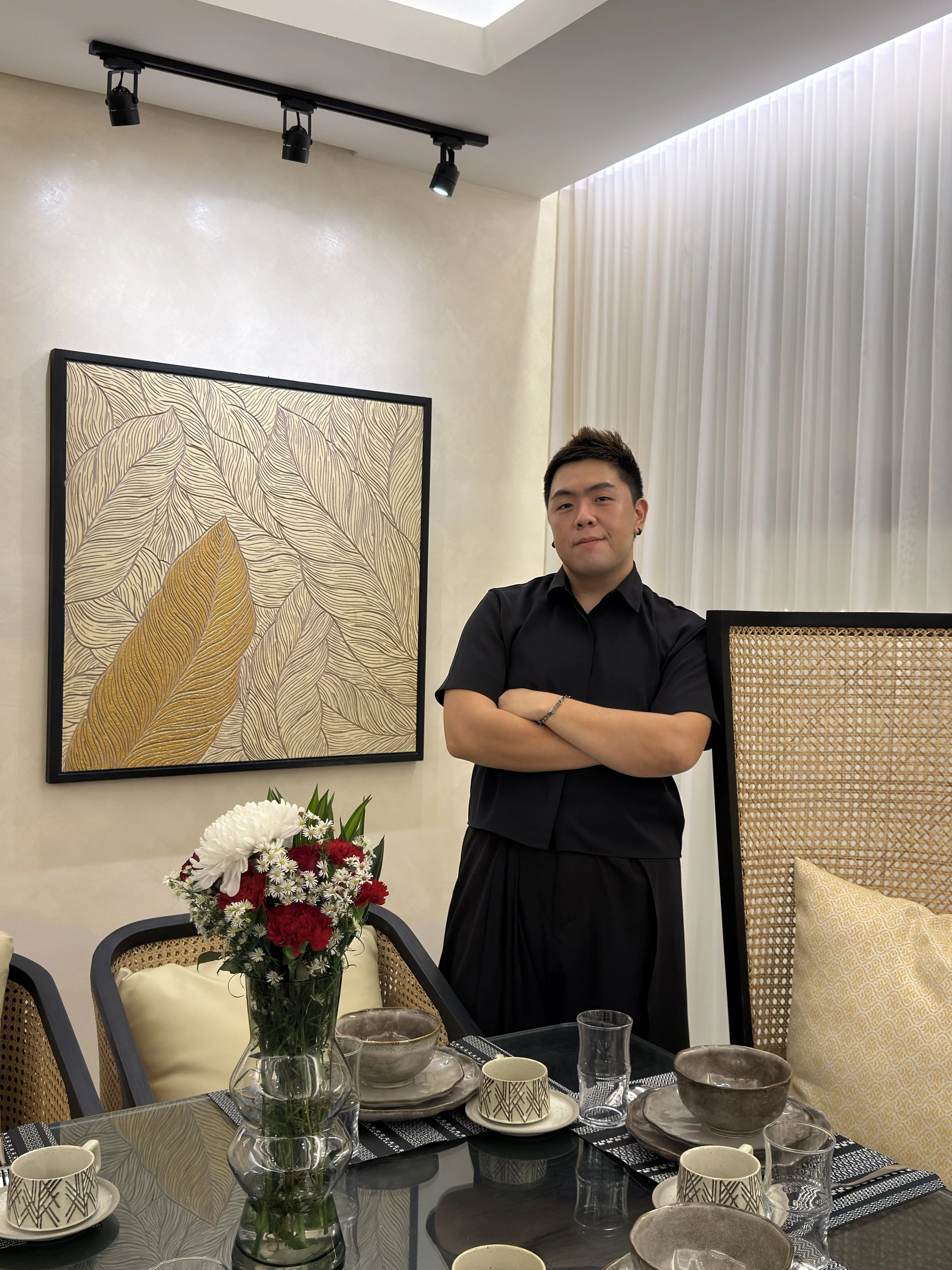 A man dressed in black standing with arms crossed in a dining room, surrounded by a table set with bowls, cups, and glasses, a vase of red and white flowers, artwork featuring abstract leaves on the wall, and a beige-colored decorative screen.