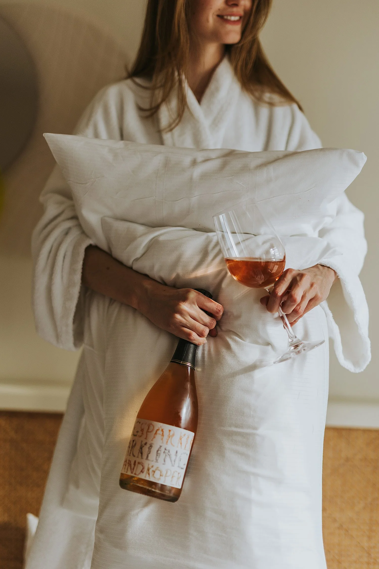 Woman in a white robe holding a pillow, a bottle of rosé wine, and a glass of rosé wine.