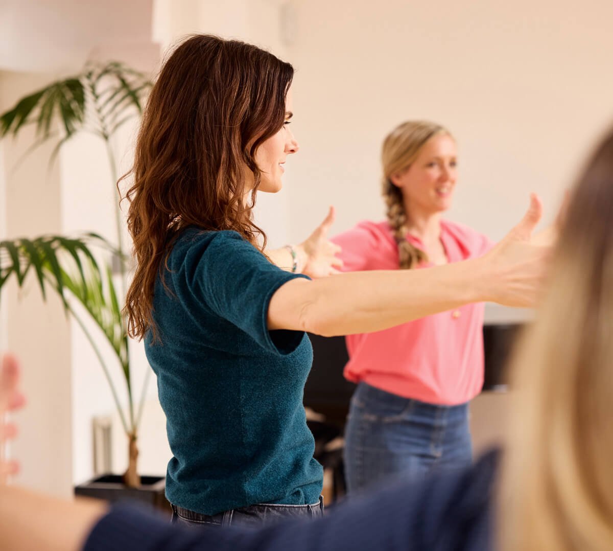 A woman with brown wavy hair wearing a navy blue shirt is leading a group exercise or dance class, with her arms outstretched and smiling at the participants. A woman with blonde hair and a pink shirt, standing in the background, is also smiling and participating.
