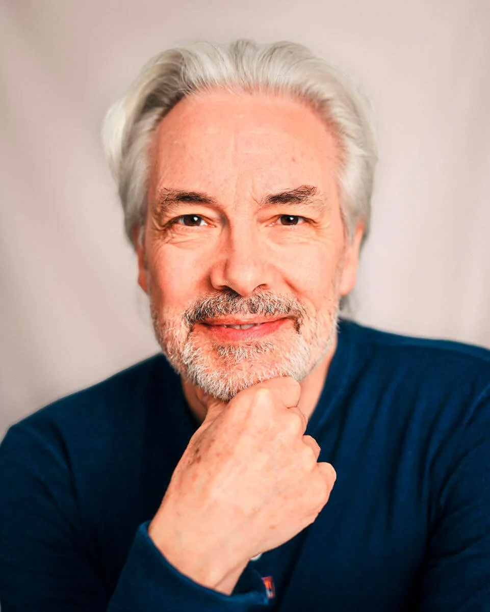 Close-up portrait of a man with white hair and a beard, smiling gently with his hand resting on his chin, wearing a dark blue shirt against a light background.