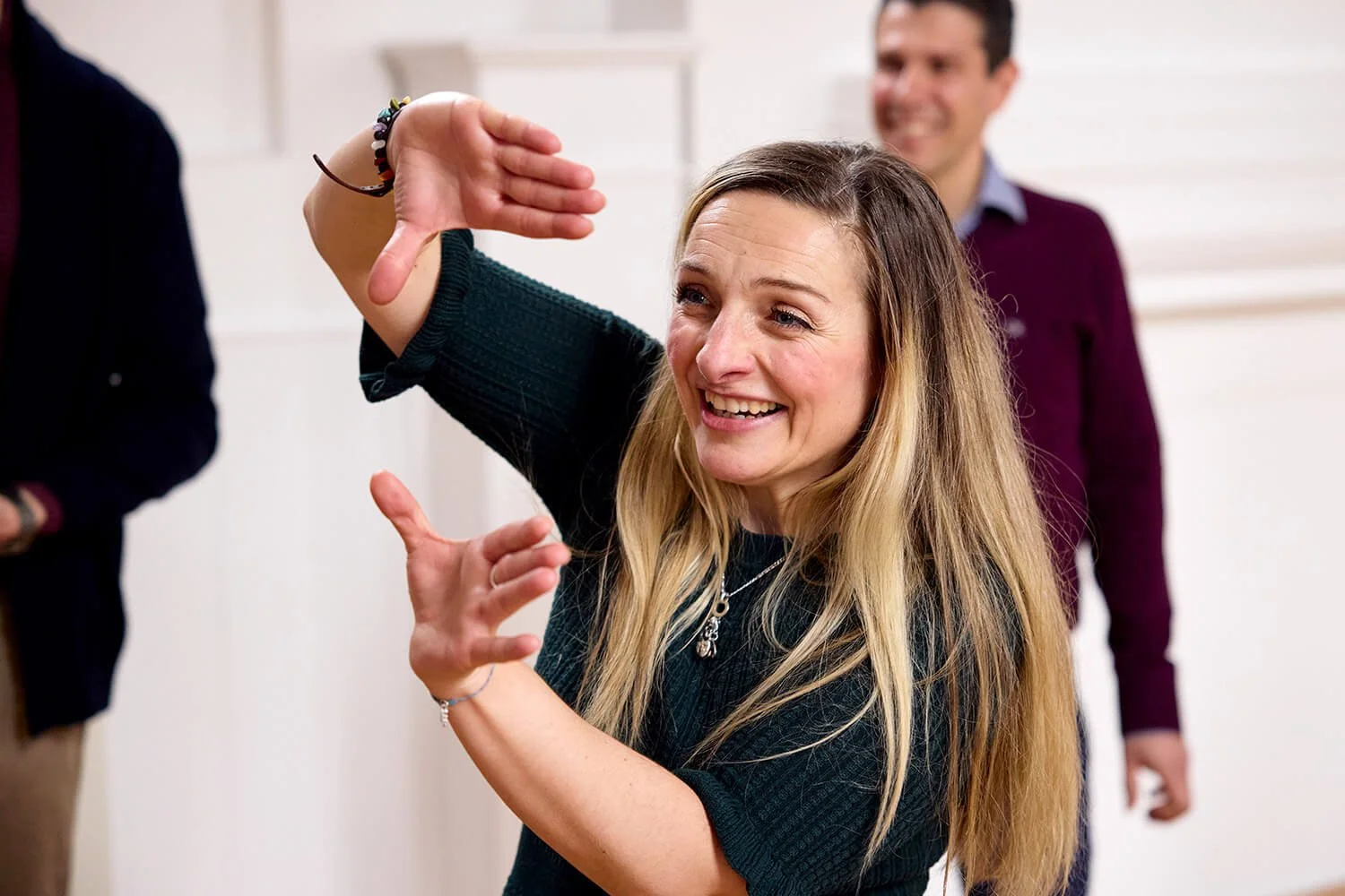 A woman with long blonde hair smiling and gesturing with her hands, with two men in the background.