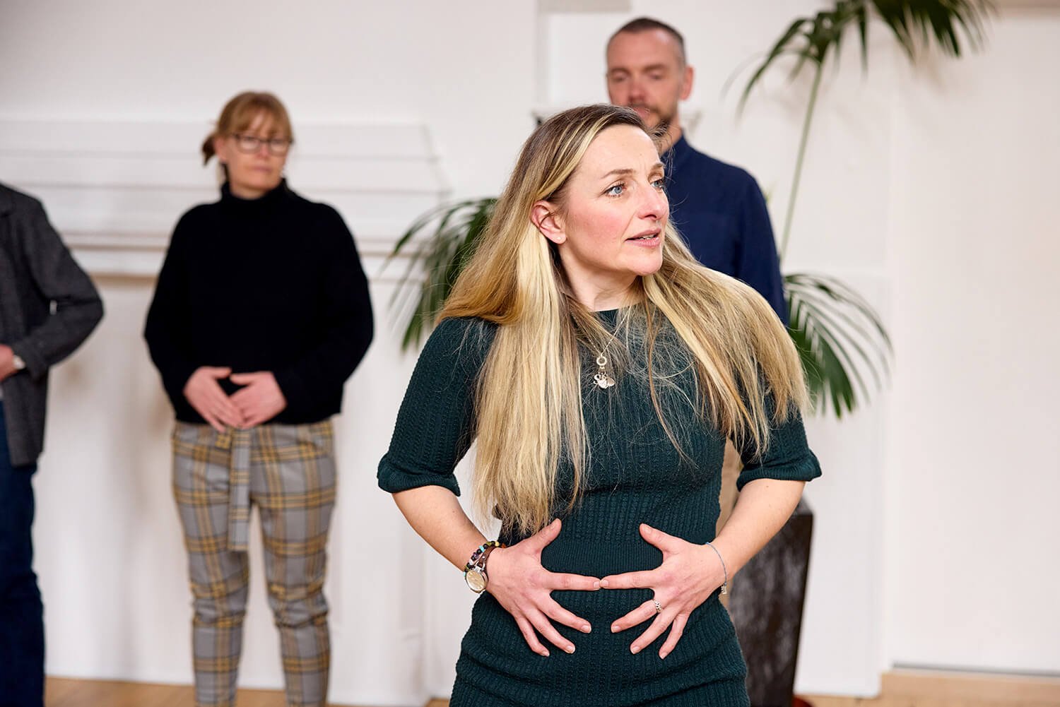A woman with long blonde hair, wearing a dark green dress and jewelry, stands with a passionate expression, hands on her stomach, in a room with other people and a large plant.