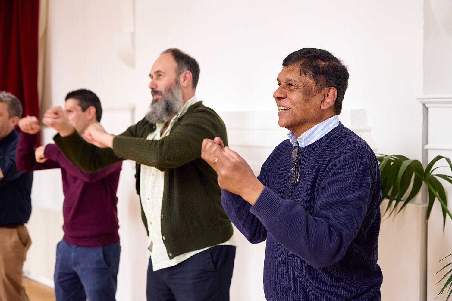 A diverse group of men standing in a line, smiling and clapping during a gathering or celebration in a bright, decorated room.