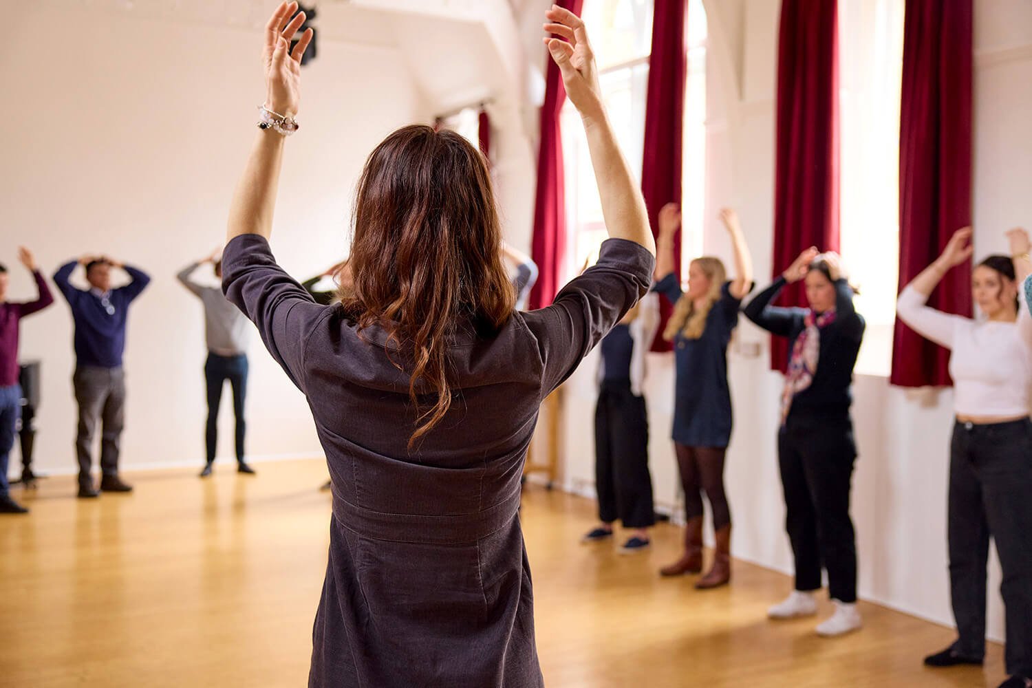 A woman with brown hair leading a movement exercise in a room with red curtains and natural light, with participants following her movements.