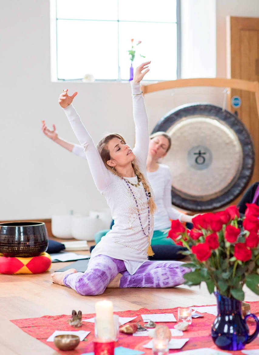 Women practicing yoga or meditation in a bright room with a gong in the background, candles, flowers, and ritual items on a red cloth.