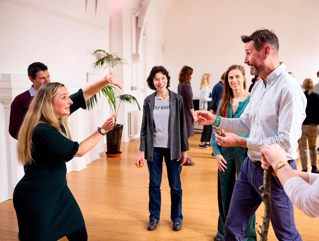 Group of people socializing in a bright room, with a woman gesturing towards a man who is smiling and holding a small object.