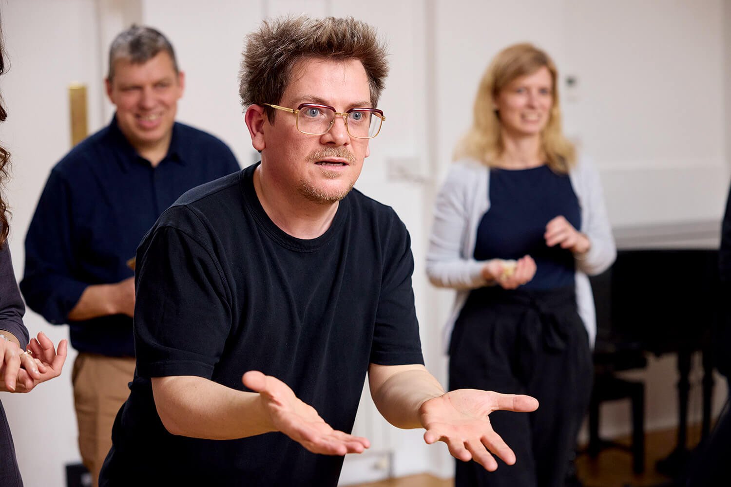 A man with glasses and a curious expression gestures with his hands while a group of people, including a woman and a man, watch during a discussion in a room.