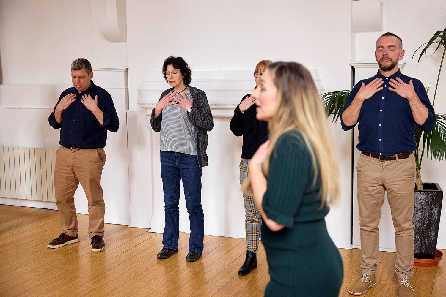 Five people standing indoors with eyes closed and hands on their chests, practicing a breathing or meditation exercise. They are arranged in a row with a woman in the front slightly out of focus, and a man on each end in the background.