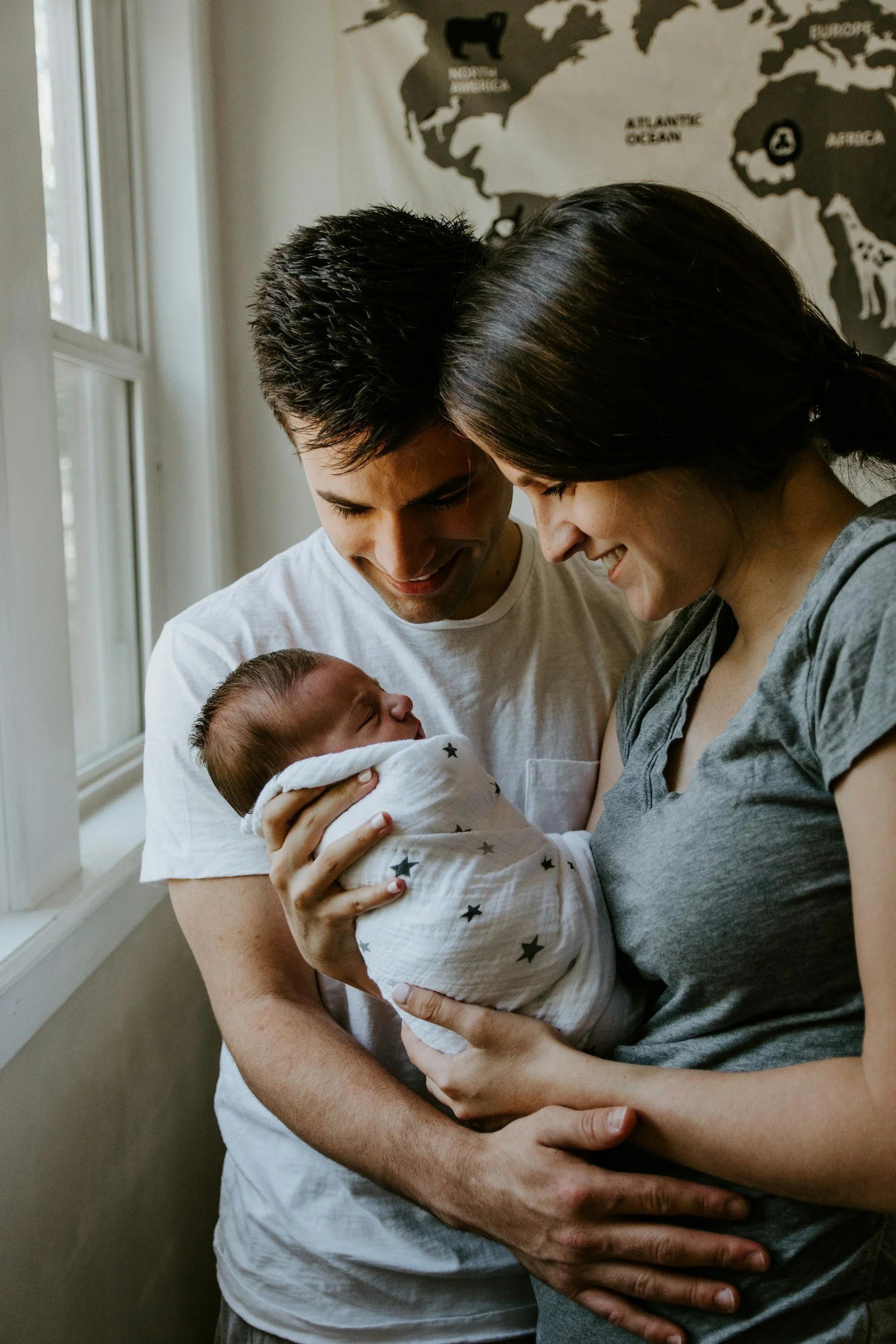 A happy family of three, including a newborn baby wrapped in a star-patterned blanket, is gathered indoors near a window with a world map on the wall behind them, smiling and looking at the baby.