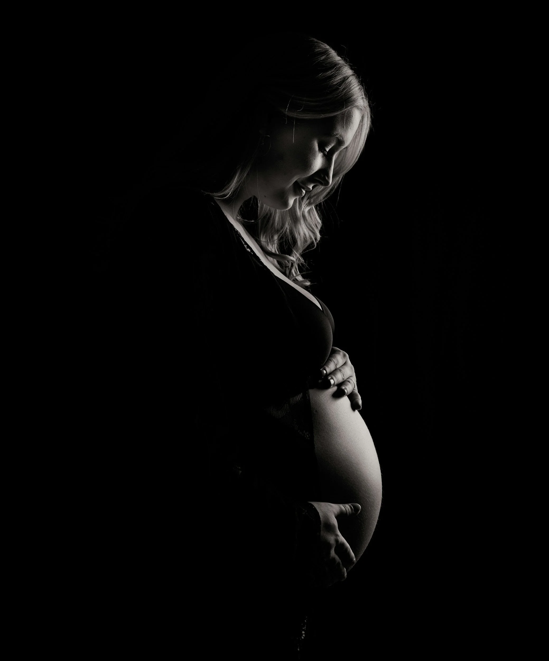 A black and white photo of a pregnant woman smiling and gently touching her belly, with her head bowed and eyes closed, against a dark background.