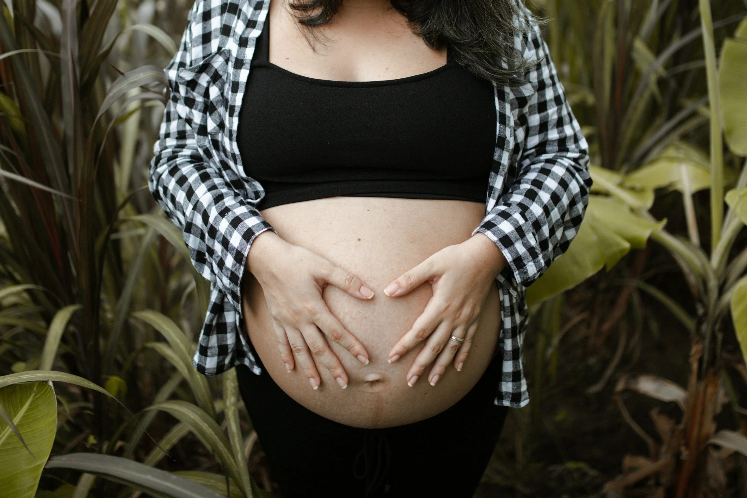 Close-up of pregnant woman outdoors, holding her belly with both hands, surrounded by plants.