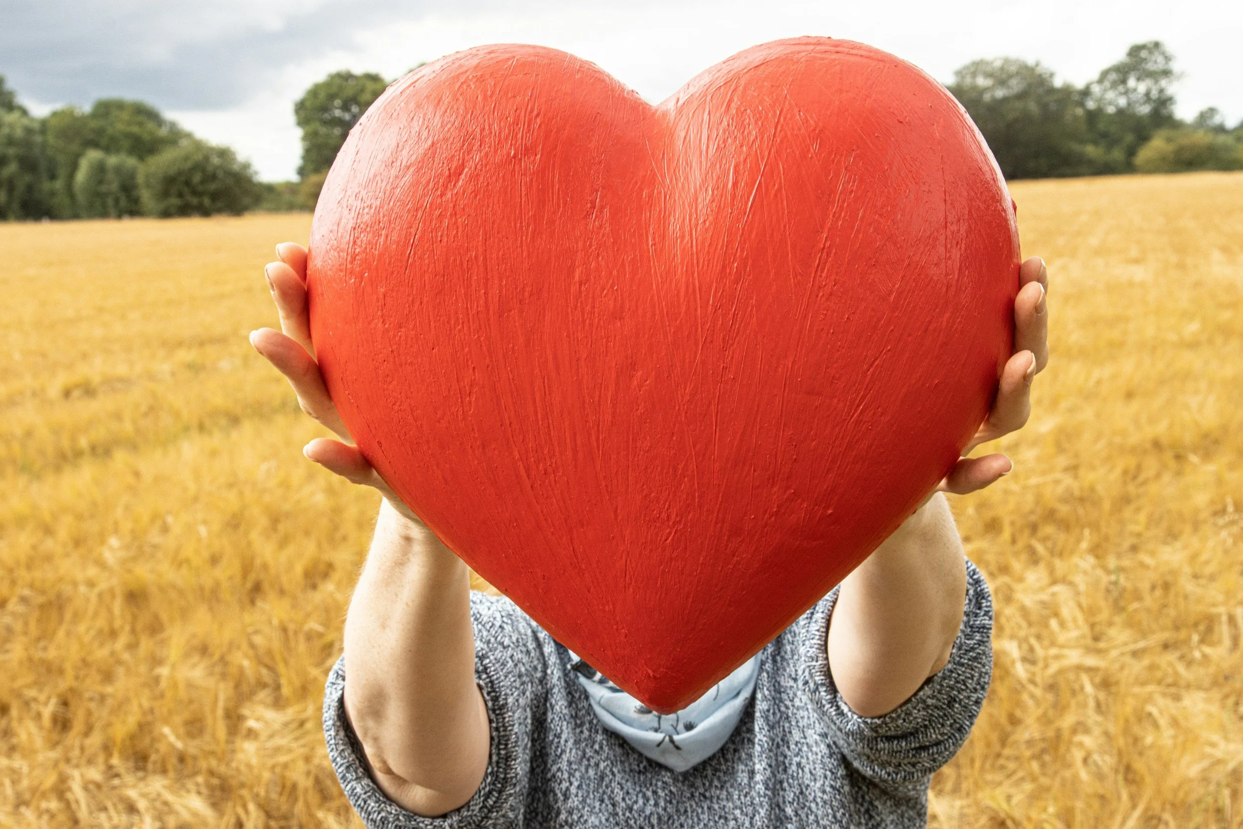 Child holding a large red heart-shaped object in front of their face in an outdoor field with trees in the background.