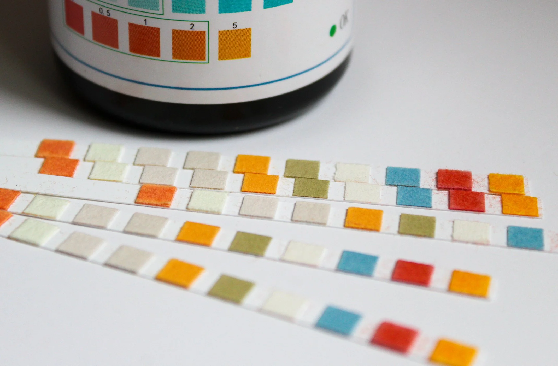 Close-up of colored pH test strips laid out on a white surface, with a partially visible pH testing meter in the background.