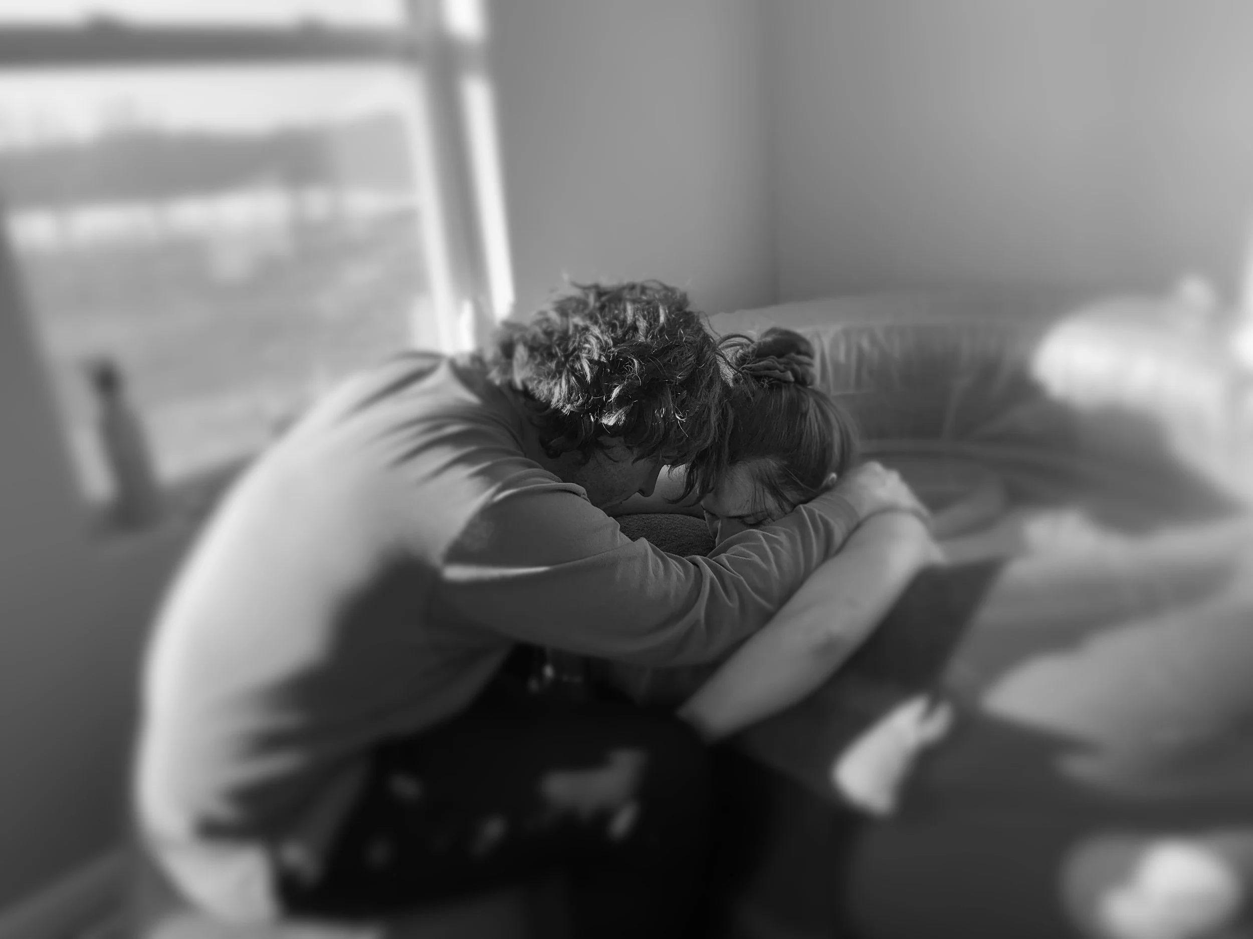 Two people embracing on a bed inside a room with sunlight coming through the window.