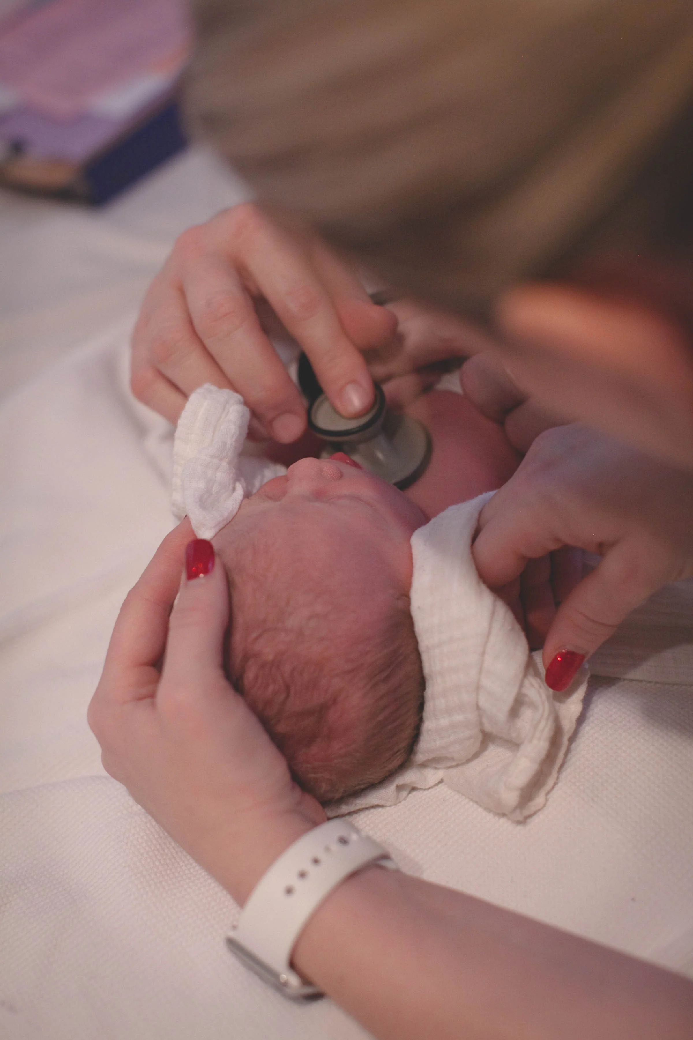 A newborn baby being examined with a stethoscope by a healthcare professional, with a woman holding the baby's head and shoulder.