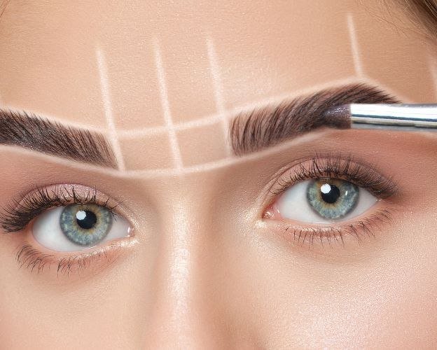 Close-up of a woman's face showing neatly groomed eyebrows. A makeup brush is being used on the right eyebrow, with white lines indicating mapping for eyebrow shaping.
