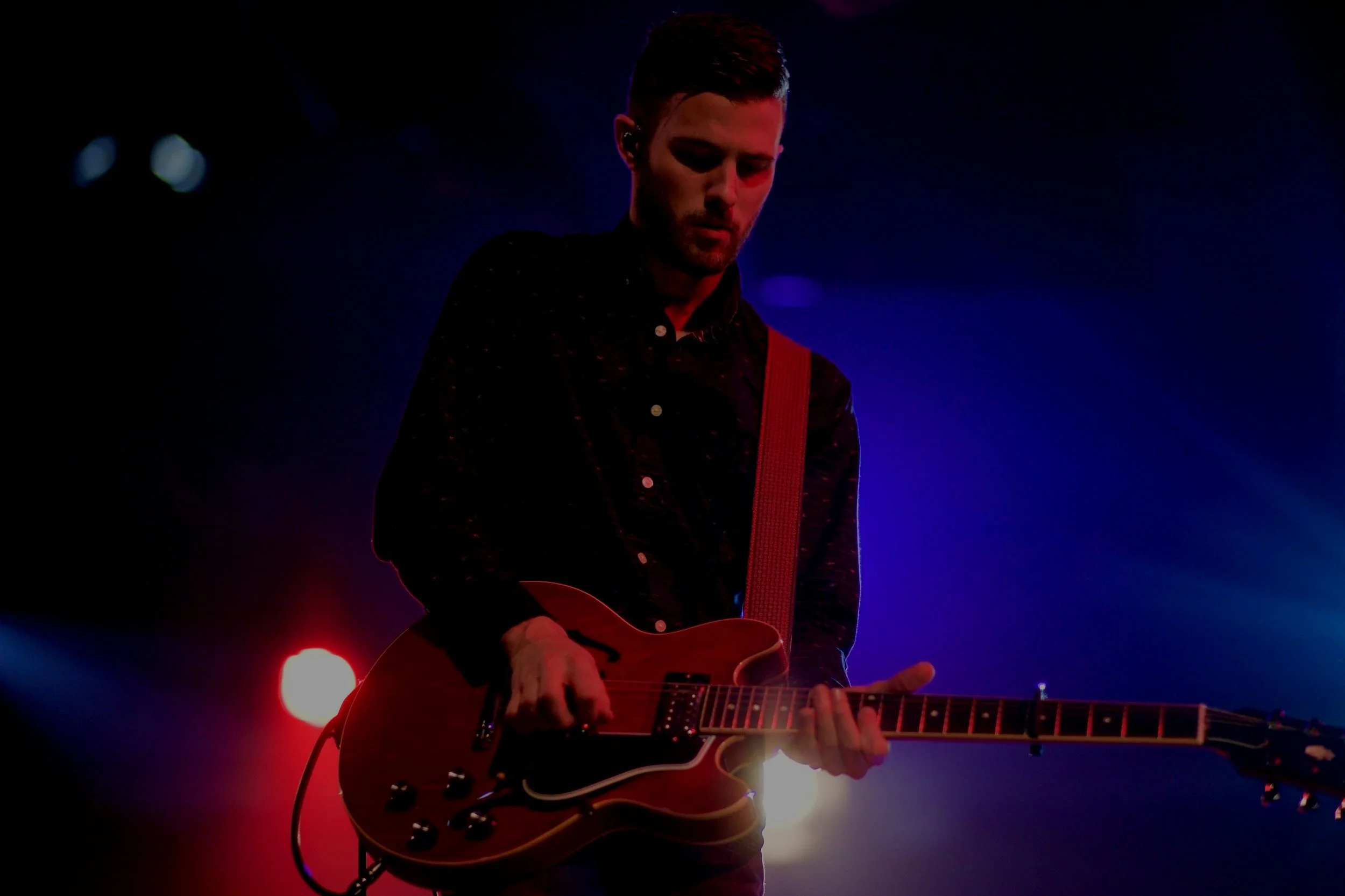 A man playing an electric guitar on stage with red and blue stage lighting.
