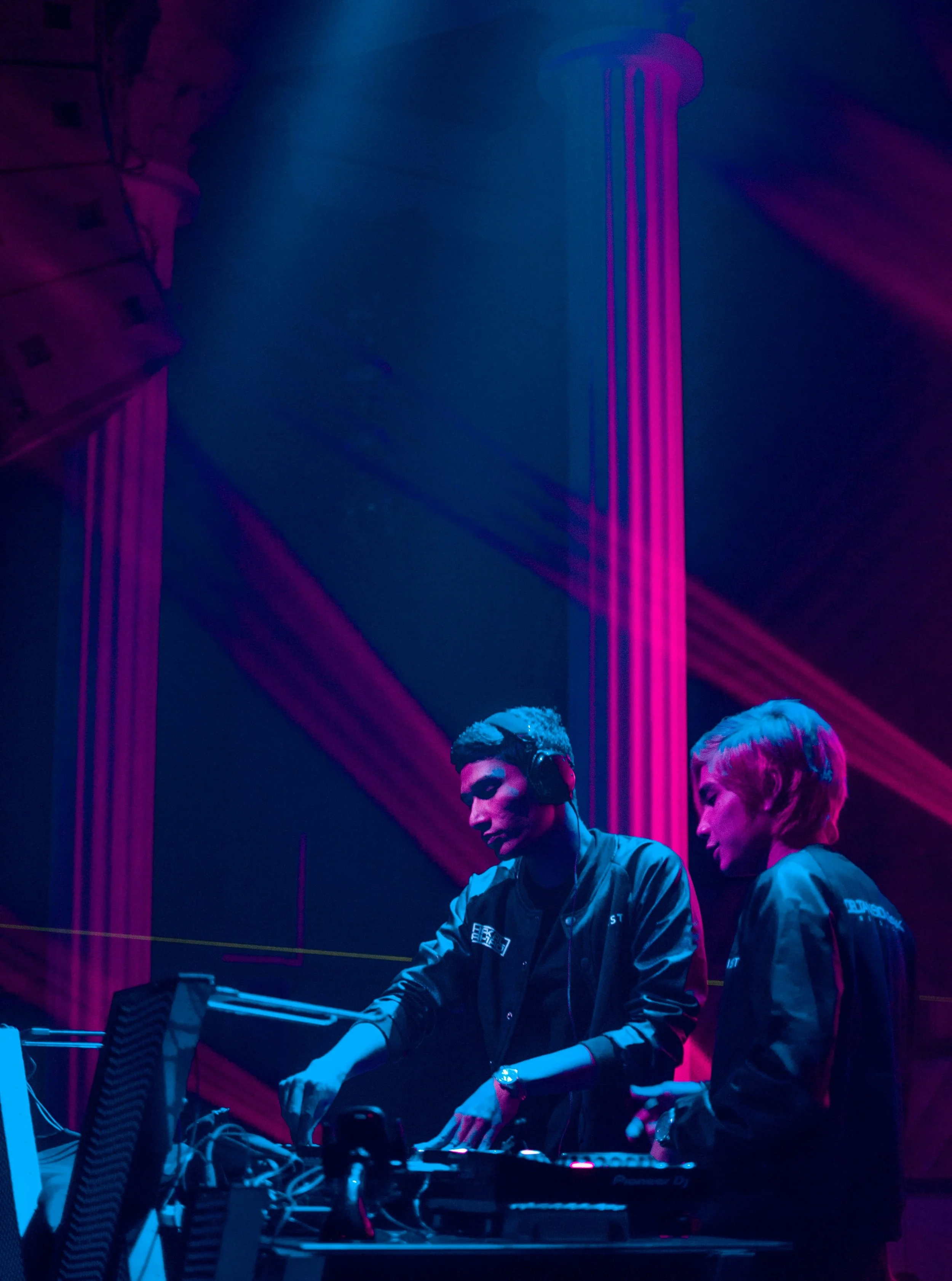 Two DJs, one male with headphones and one female, are performing on stage with colorful pink and blue lighting, and DJ equipment.
