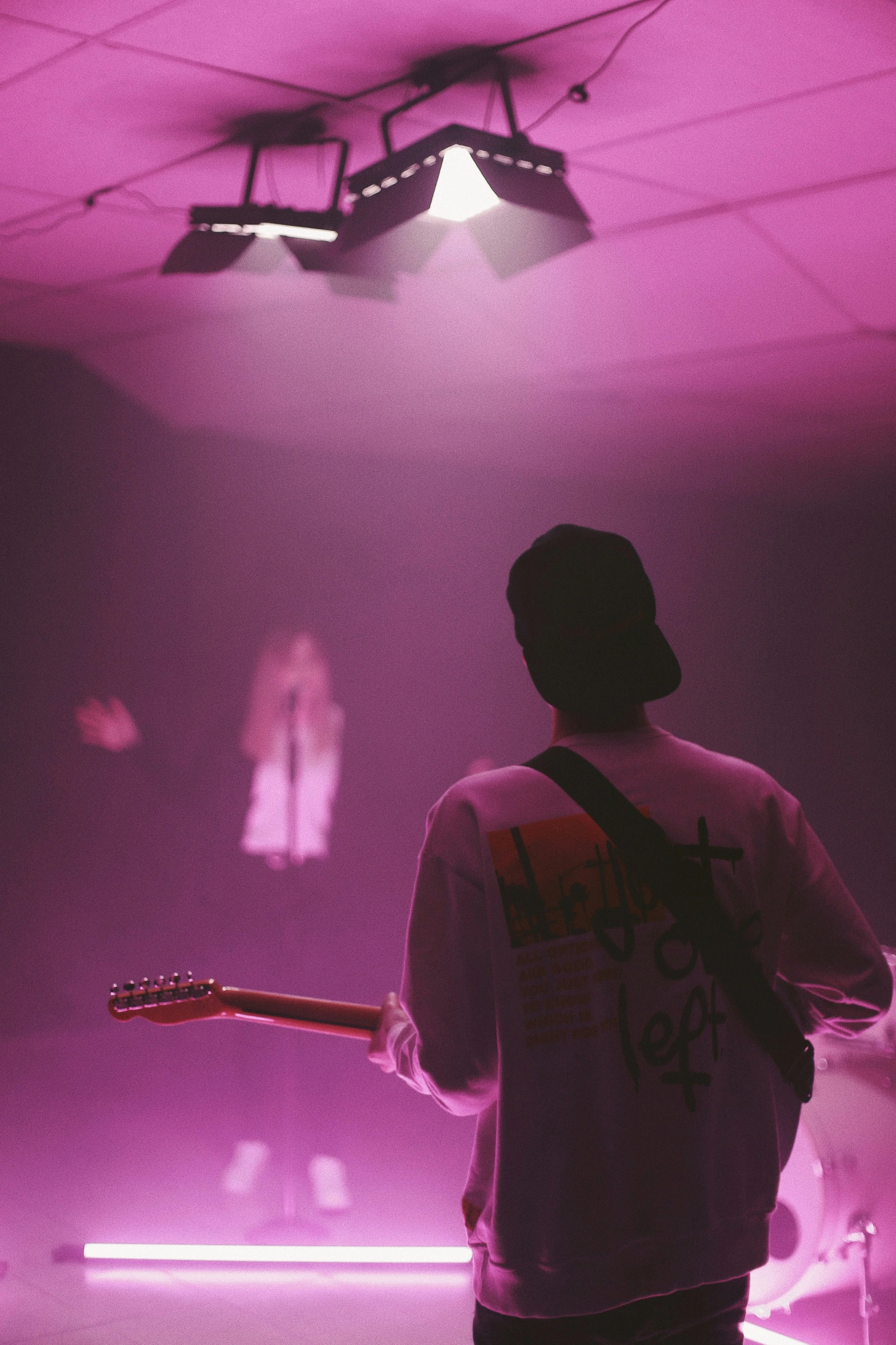 A person in a dark cap and white jacket playing an electric guitar under pink stage lighting, with a person on stage in the background singing into a microphone.