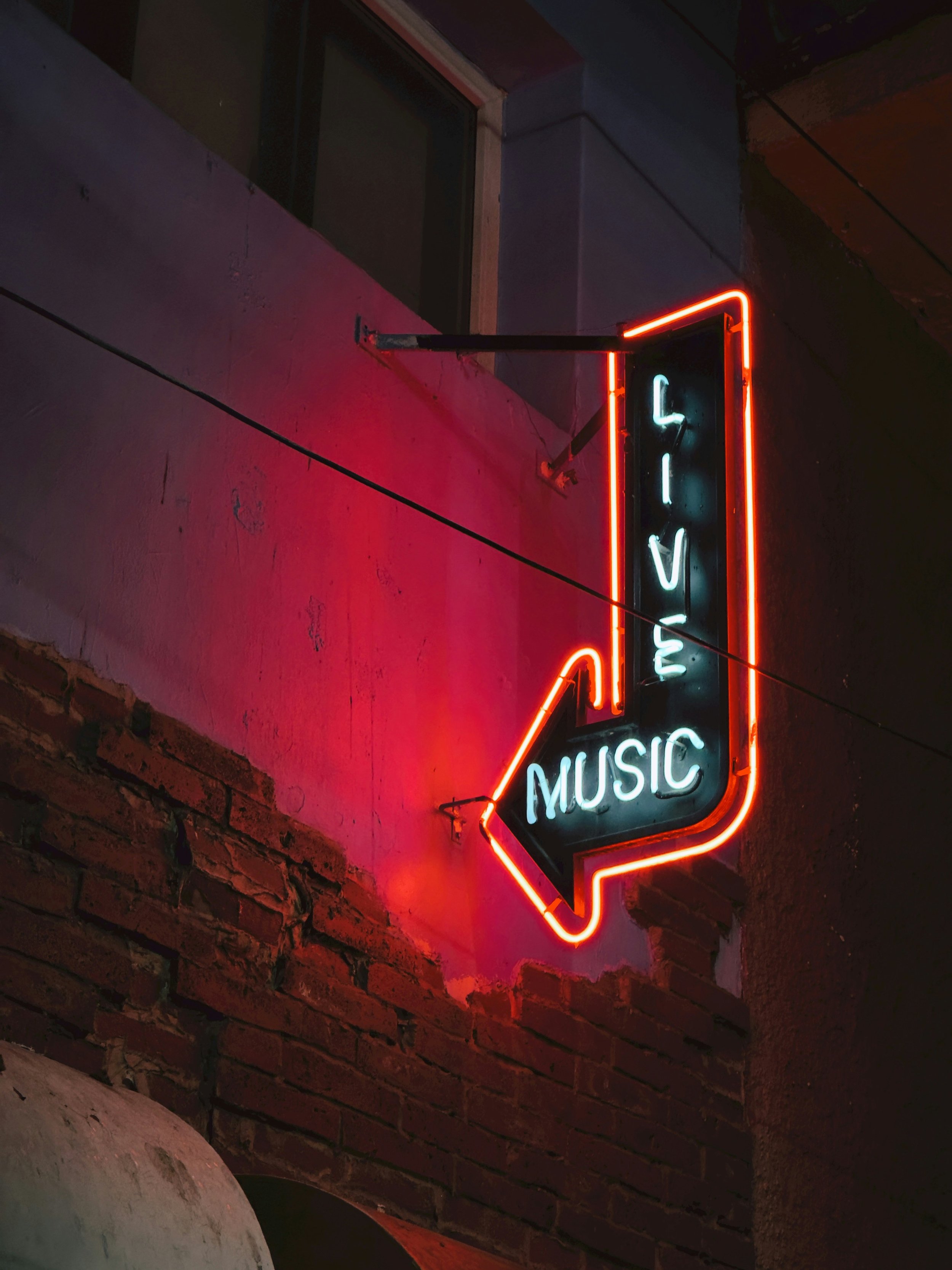 Neon sign with an arrow pointing left, displaying the words 'Live Music' against a pink wall with brick accents.
