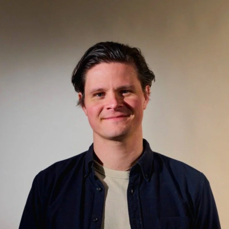 A young man with dark brown hair styled back, wearing a dark button-up shirt over a cream t-shirt, standing against a plain beige background, smiling slightly.