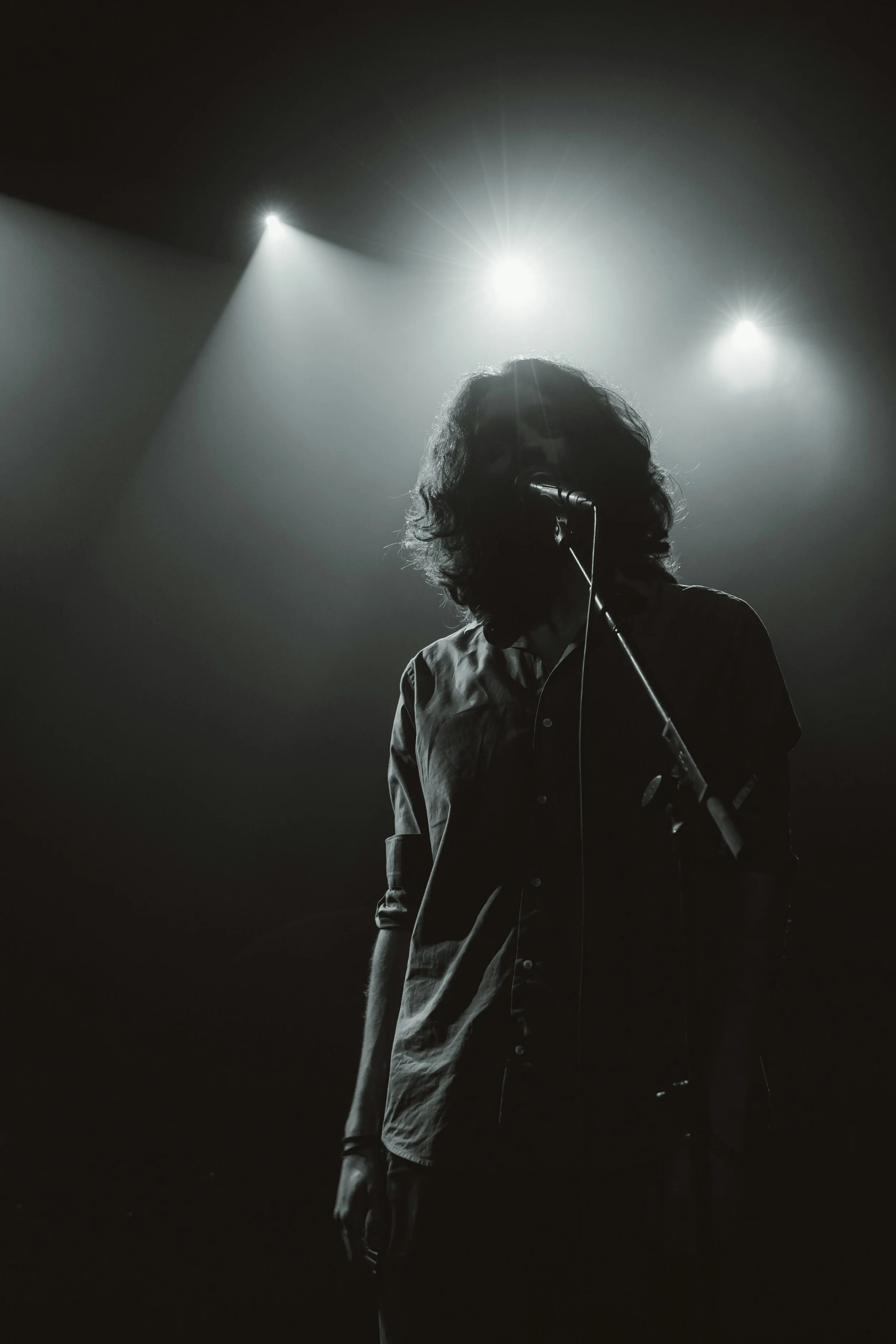 A person with curly hair performing on stage, standing in front of a microphone, with stage lights shining behind.