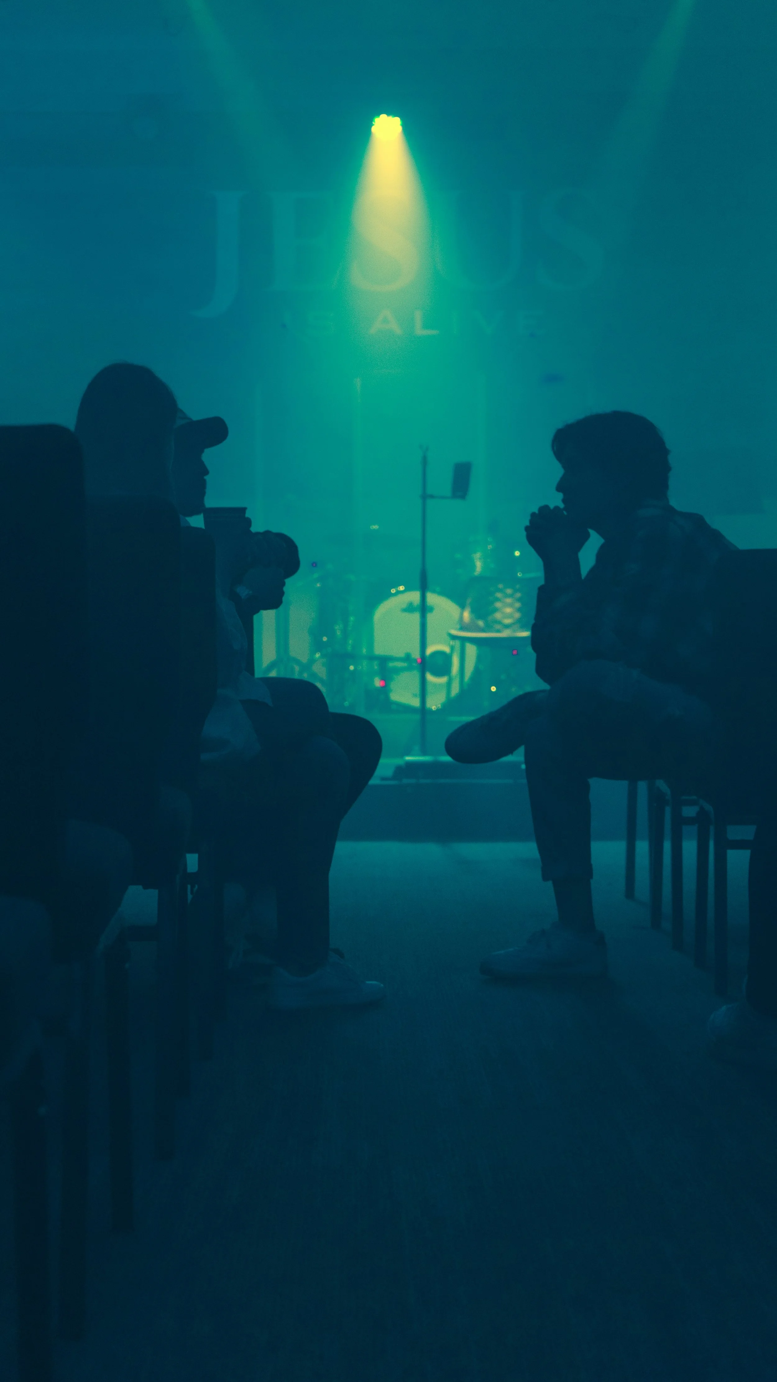 Silhouetted group of people seated in a dimly lit room with a stage and large illuminated sign reading 'JESUS' in the background.