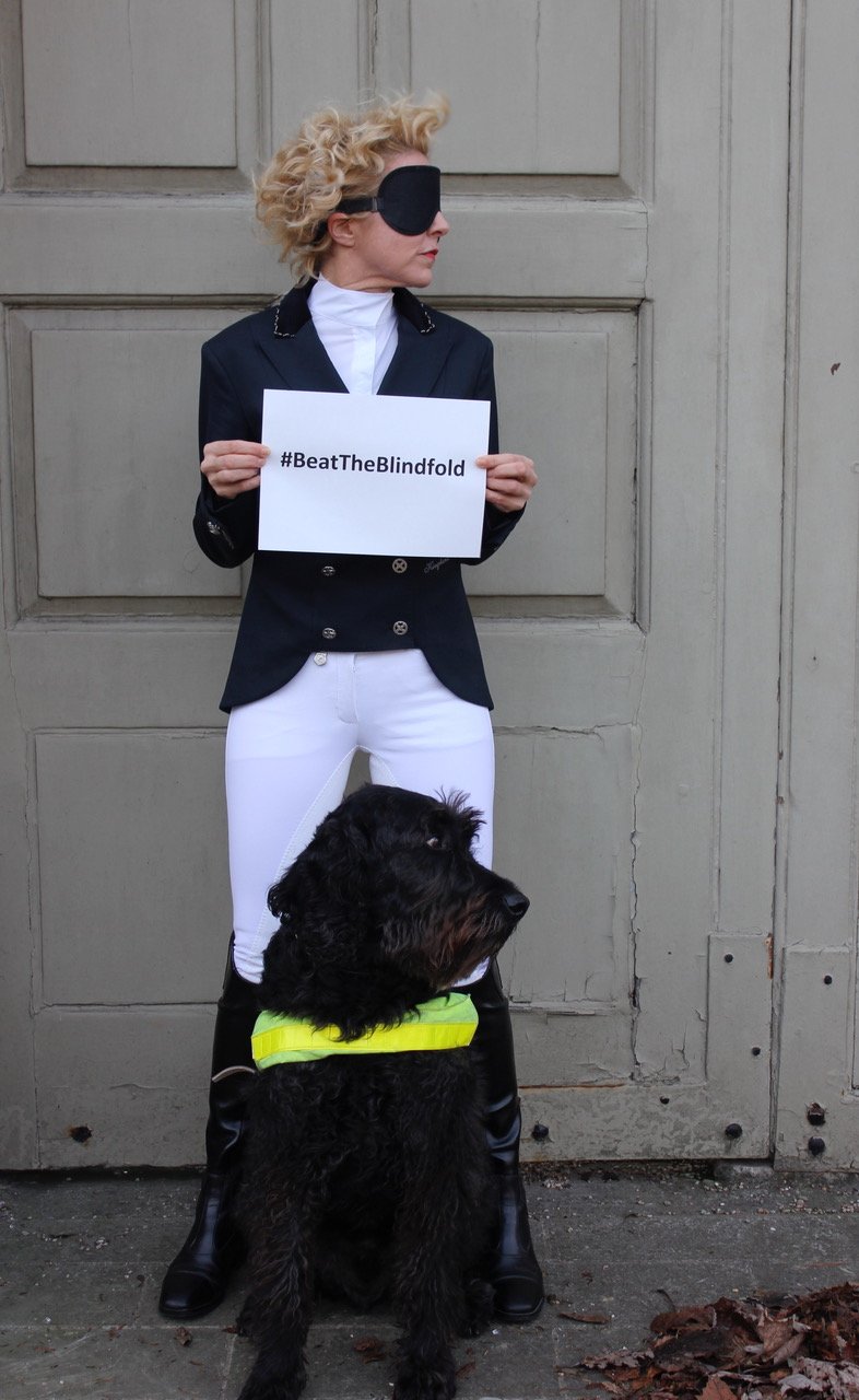 Verity Smith in equestrian outfit standing in the street wearing a blindfold, holding a #BeatTheBlindfold sign, with her black guide dog Uffa sitting in front of her wearing a yellow harness.