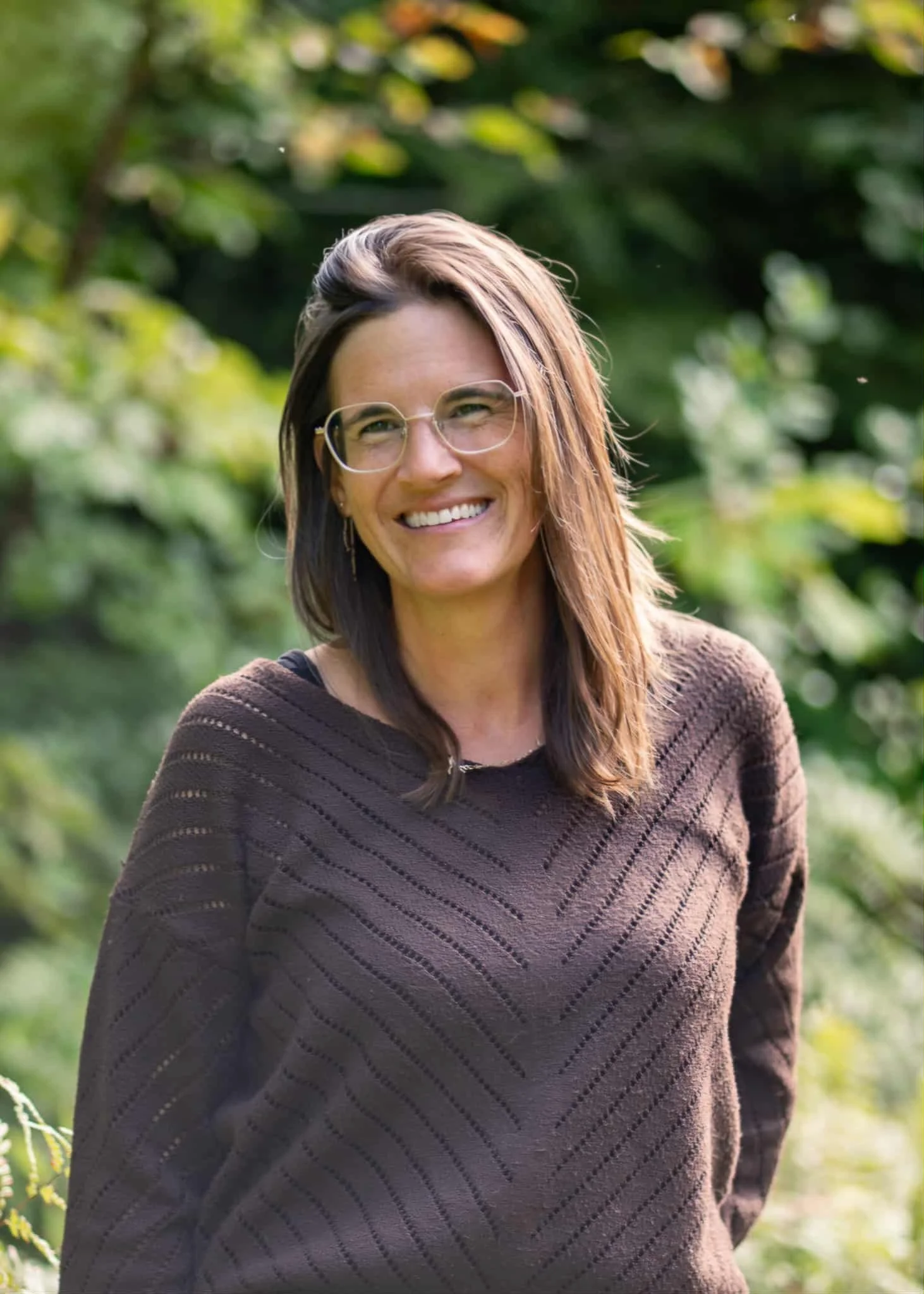 Femme souriante portant des lunettes, cheveux longs et bruns, vêtue d'un pull marron, en plein air avec un fond de nature.