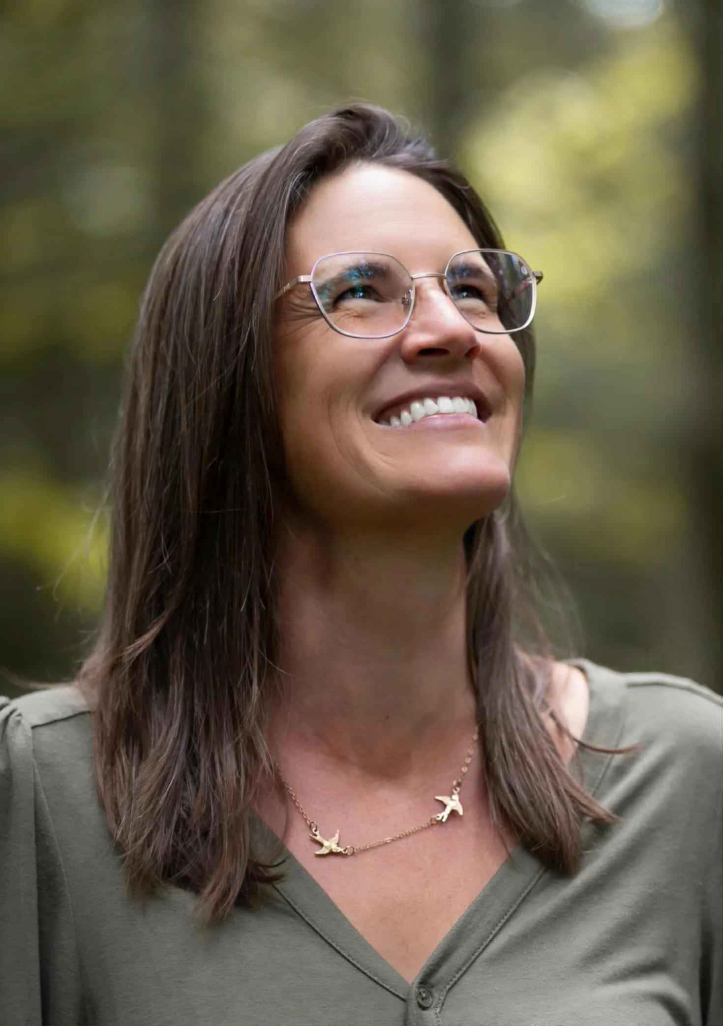 Une femme souriante portant des lunettes, une chaîne avec des oiseaux en pendentifs, et un haut gris avec une fermeture éclair.